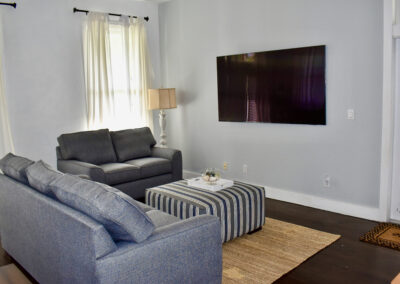 Guest cottage lounge at Saint Patrick Palace with cozy seating, striped ottoman, and mounted TV in Davie, Florida