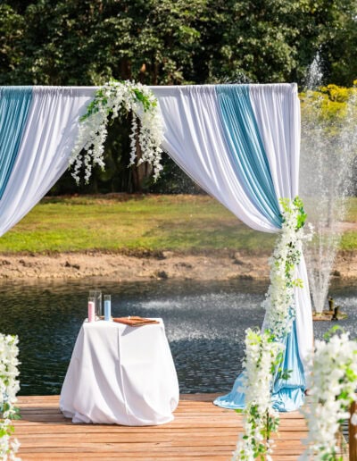 A stunning outdoor wedding ceremony setup featuring a blue and white color palette, silver chiavari chairs, and a draped arch with cascading florals, set on a wooden dock by a serene fountain. Perfect for a romantic celebration at Saint Patrick Palace in Davie, Florida.
