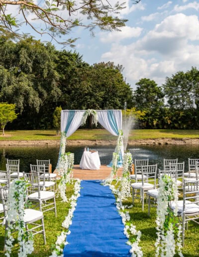 A stunning outdoor wedding ceremony setup featuring a blue and white color palette, silver chiavari chairs, and a draped arch with cascading florals, set on a wooden dock by a serene fountain. Perfect for a romantic celebration at Saint Patrick Palace in Davie, Florida.