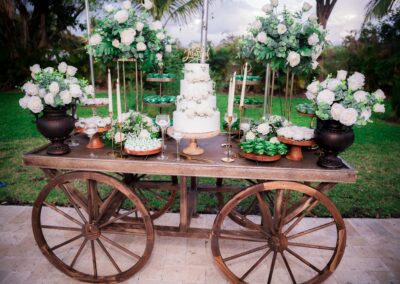 Elegant outdoor wedding dessert cart with white cake, lush florals & sweet treats.