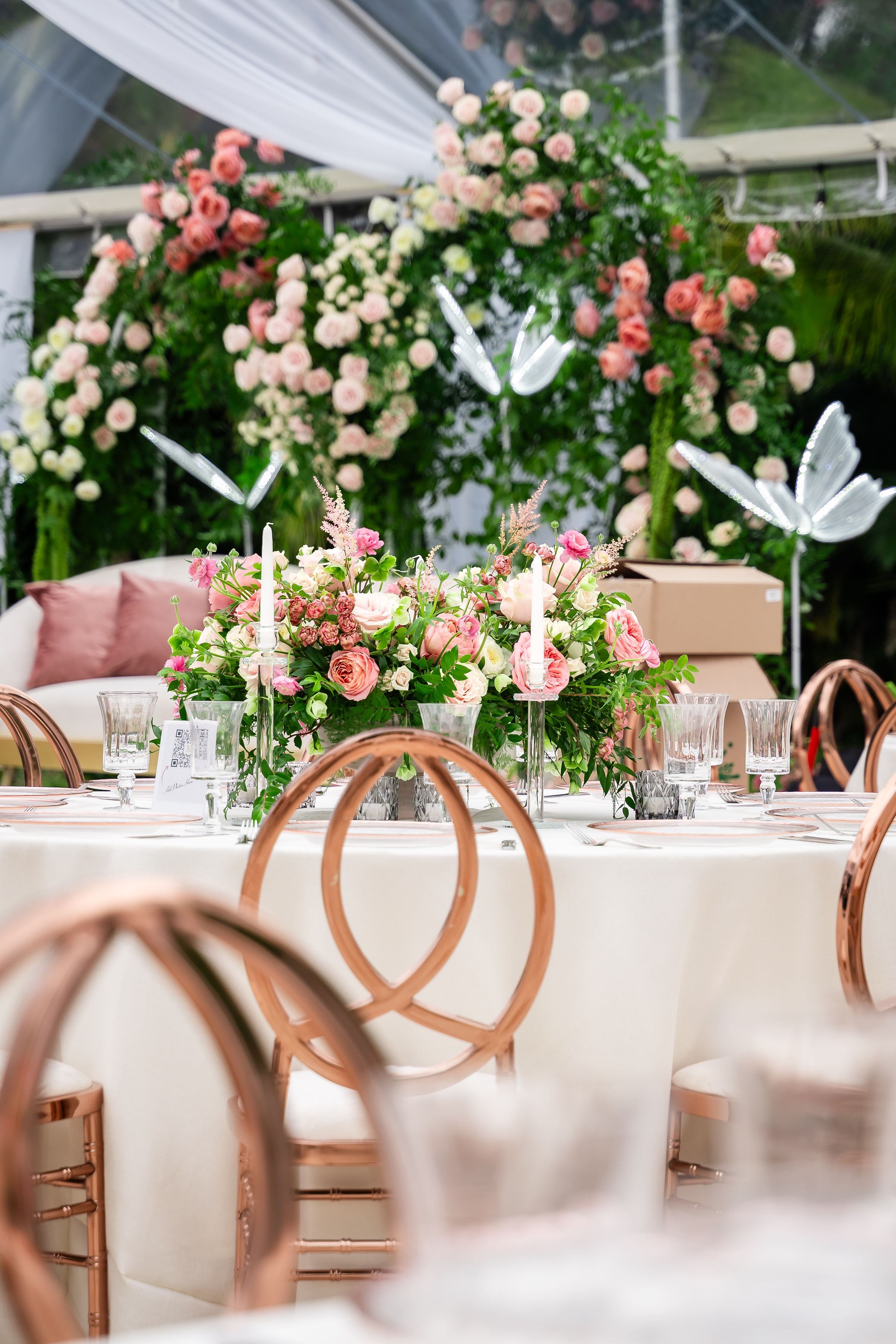 Rose gold quinceañera tablescape with roses, greenery, taper candles, and metallic accent chairs.