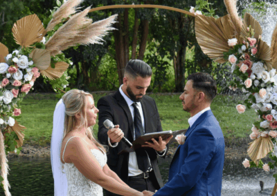 Close up of a bride and groom exchanging vows in front of a boho-style floral arch with pampas grass and a fountain background.