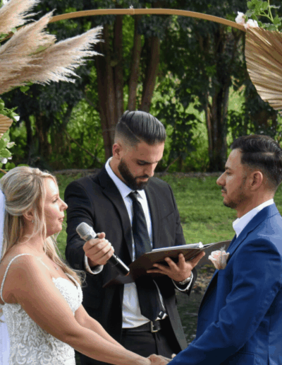 Close up of a bride and groom exchanging vows in front of a boho-style floral arch with pampas grass and a fountain background.