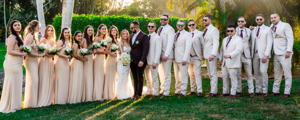 Large bridal party in beige dresses and suits posing on the manicured lawns of Saint Patrick Palace.