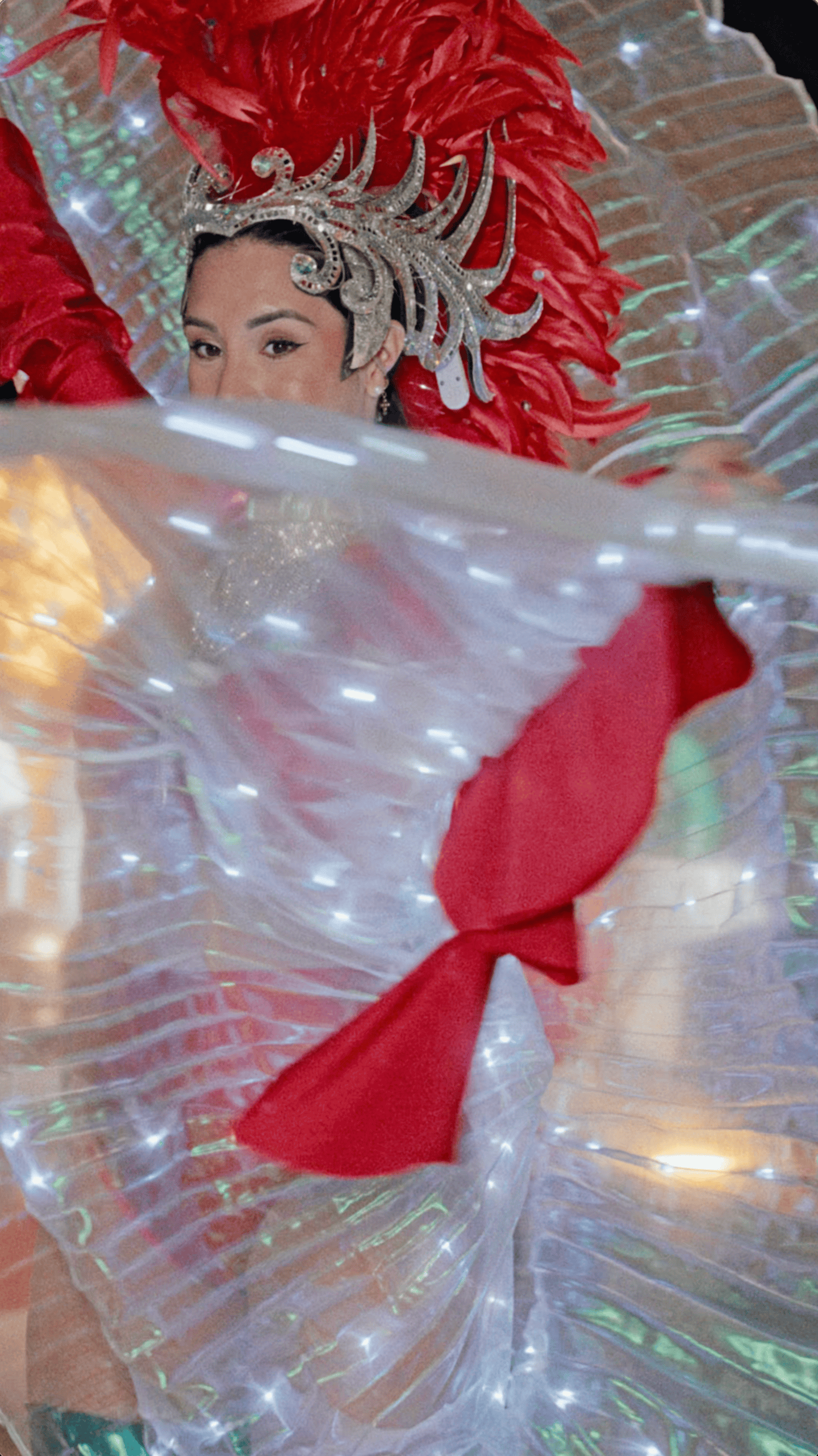 Holiday performer wearing a red feathered headdress and glowing illuminated wings during an evening celebration.
