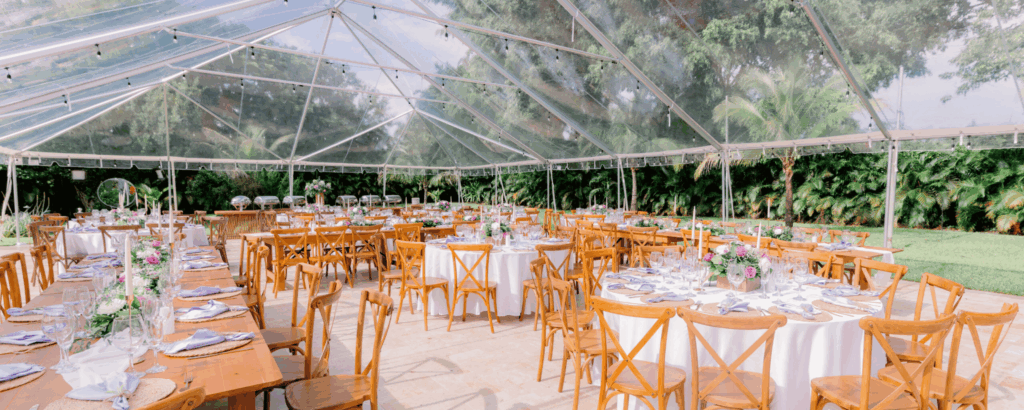 Interior view of a clear top tent wedding reception with wooden cross-back chairs and farm tables during the day.