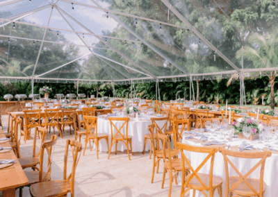 Interior view of a clear top tent wedding reception with wooden cross-back chairs and farm tables during the day.