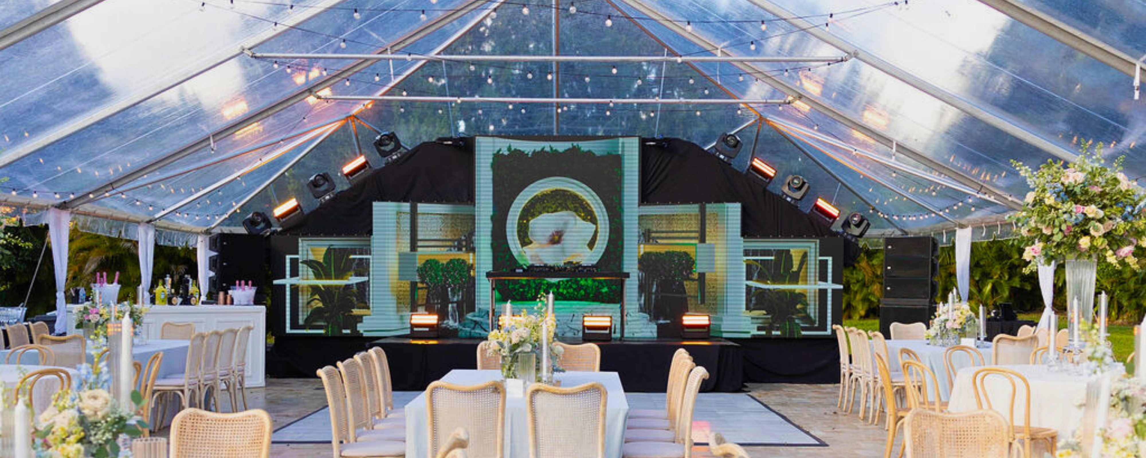 Interior of a clear-top tent reception featuring bistro lighting, LED DJ wall, and elegant table settings.