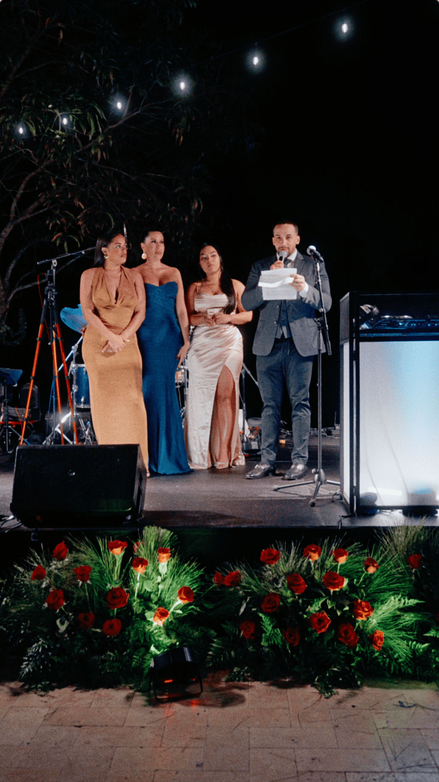 Four formally dressed guests on an outdoor stage as one delivers a speech during a holiday celebration.