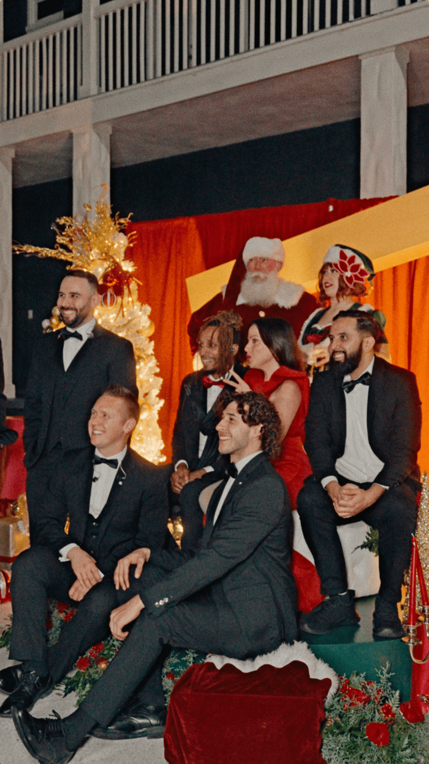 Group of holiday party guests posing with Santa Claus and a Christmas elf in front of festive decorations.