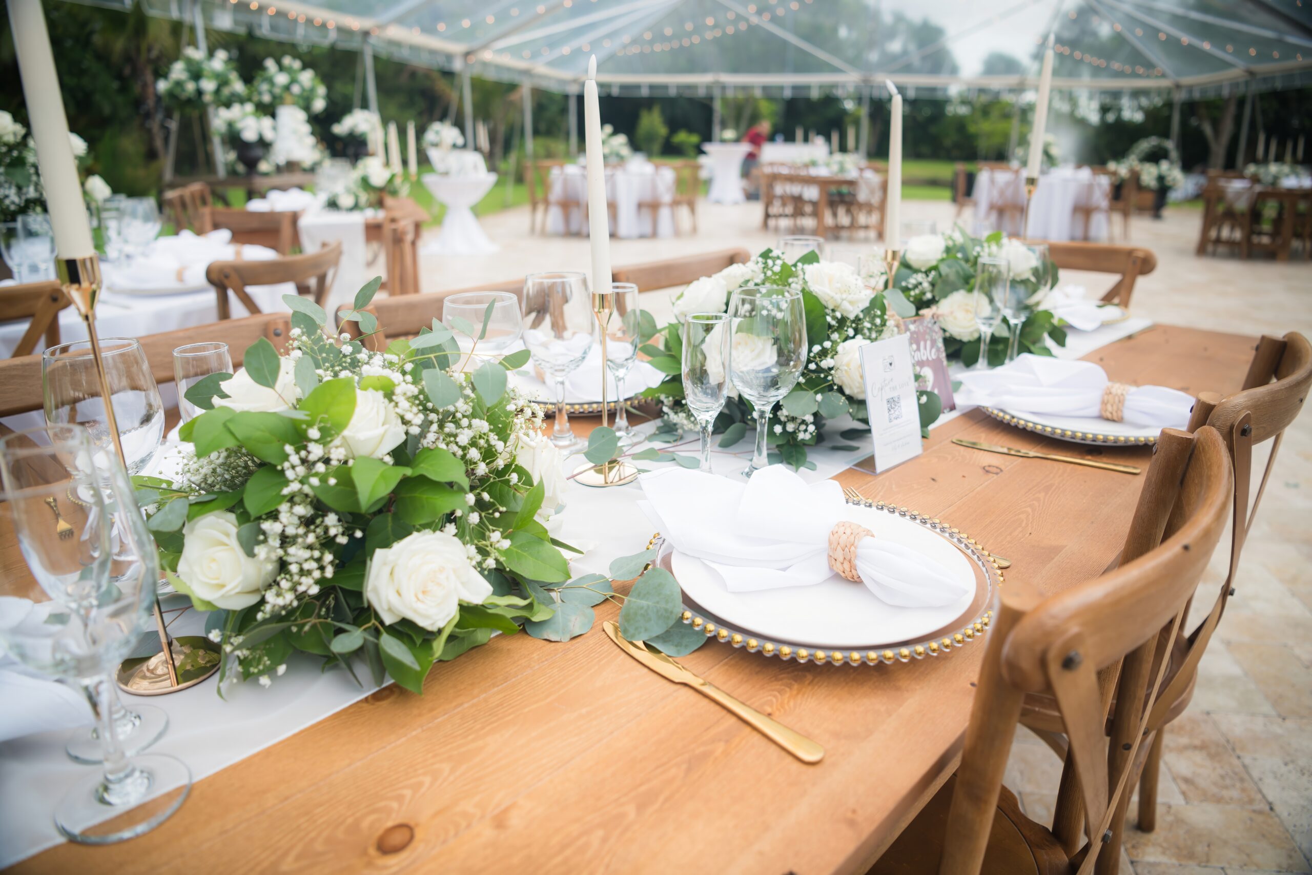 Elegant retirement party table with white roses, greenery, and gold accents under a clear tent.