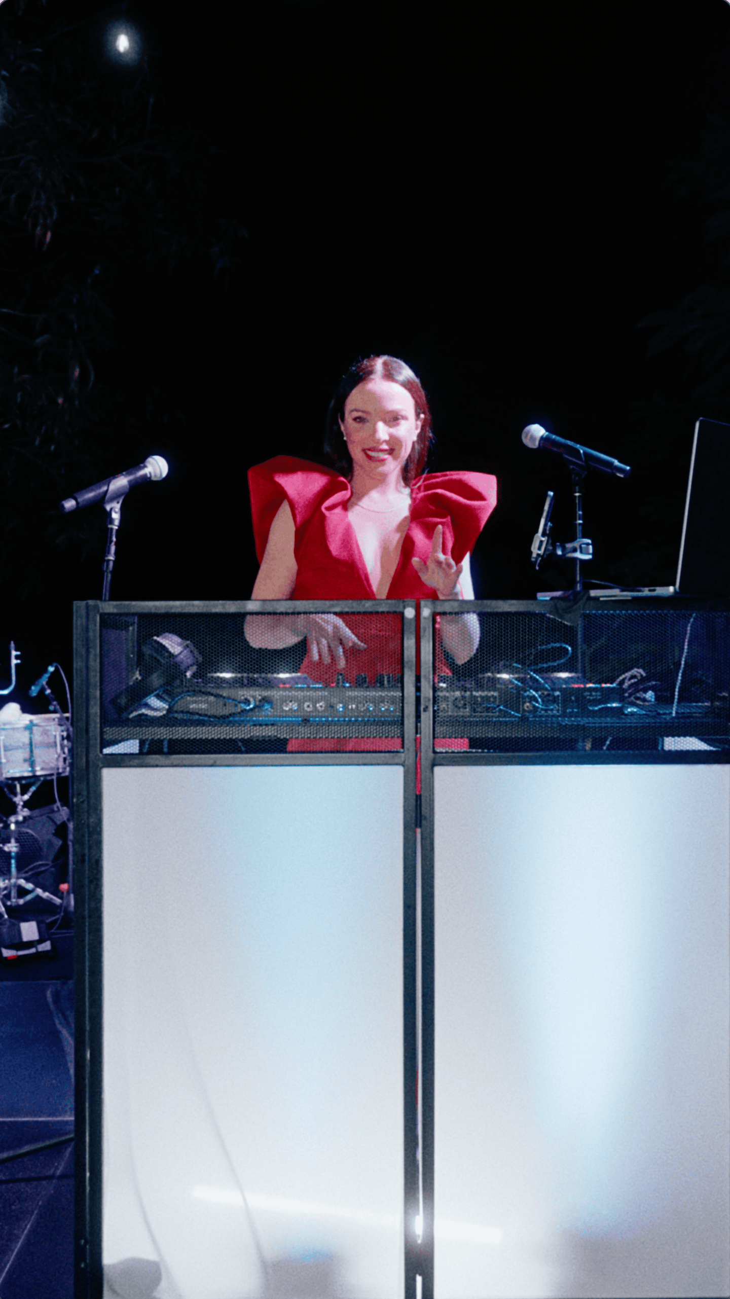 Female DJ in a red dress performing behind an illuminated DJ booth at an outdoor holiday party.
