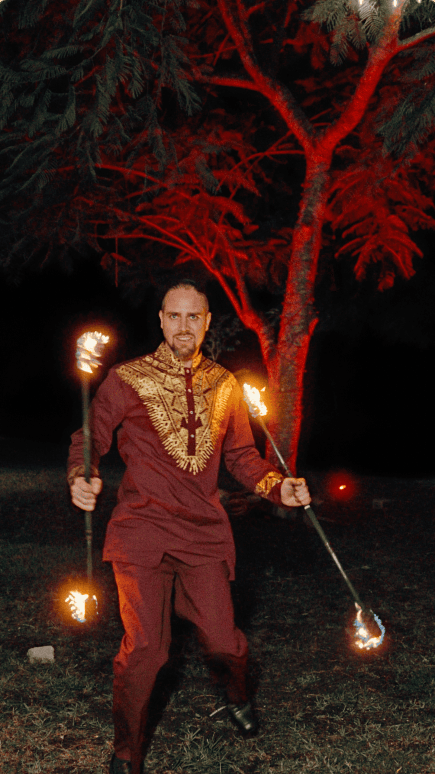 Fire performer holding two flaming staffs beneath red-lit trees during an outdoor holiday party.