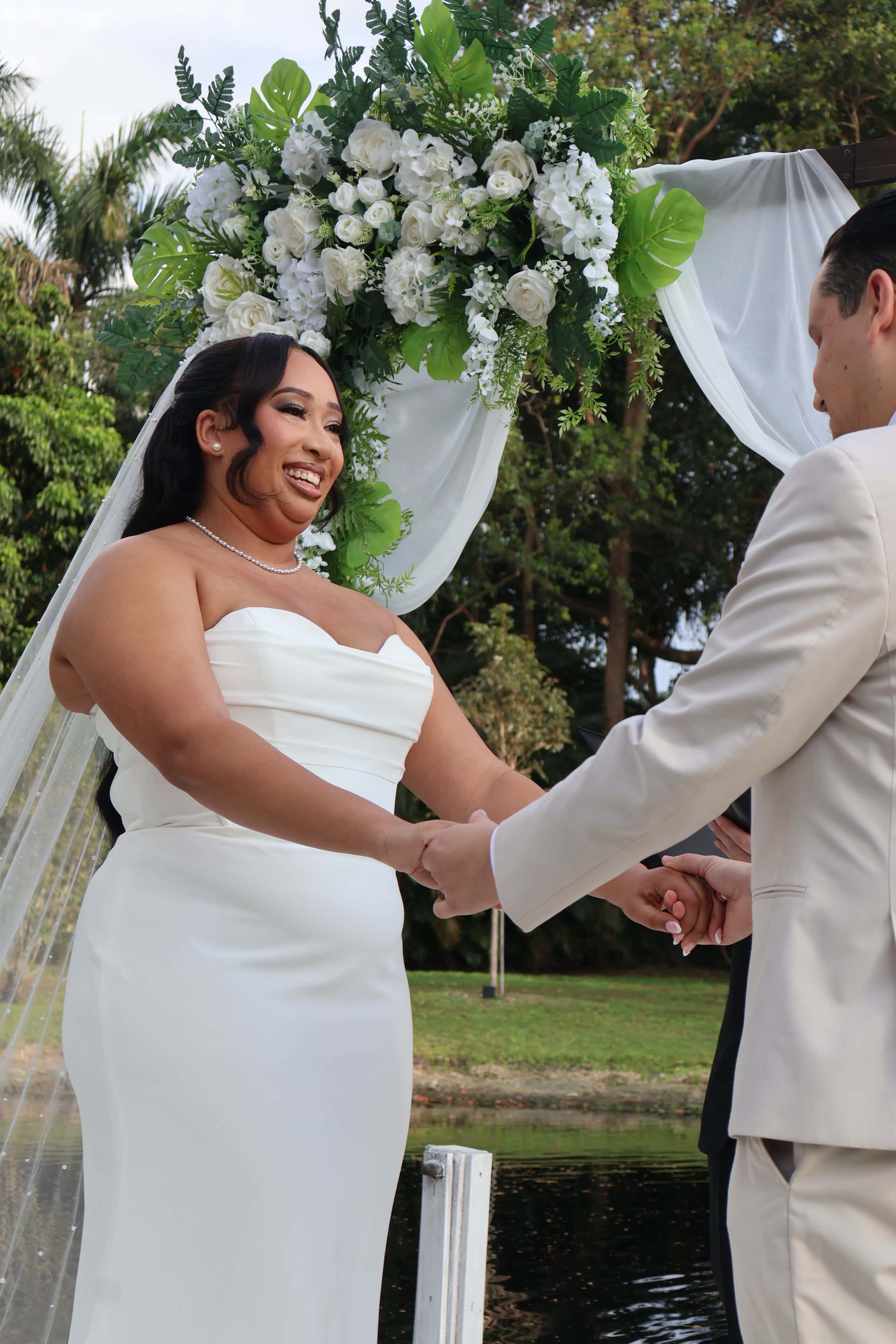 Bride and groom holding hands beneath a floral arbor during a waterfront wedding ceremony in Florida.