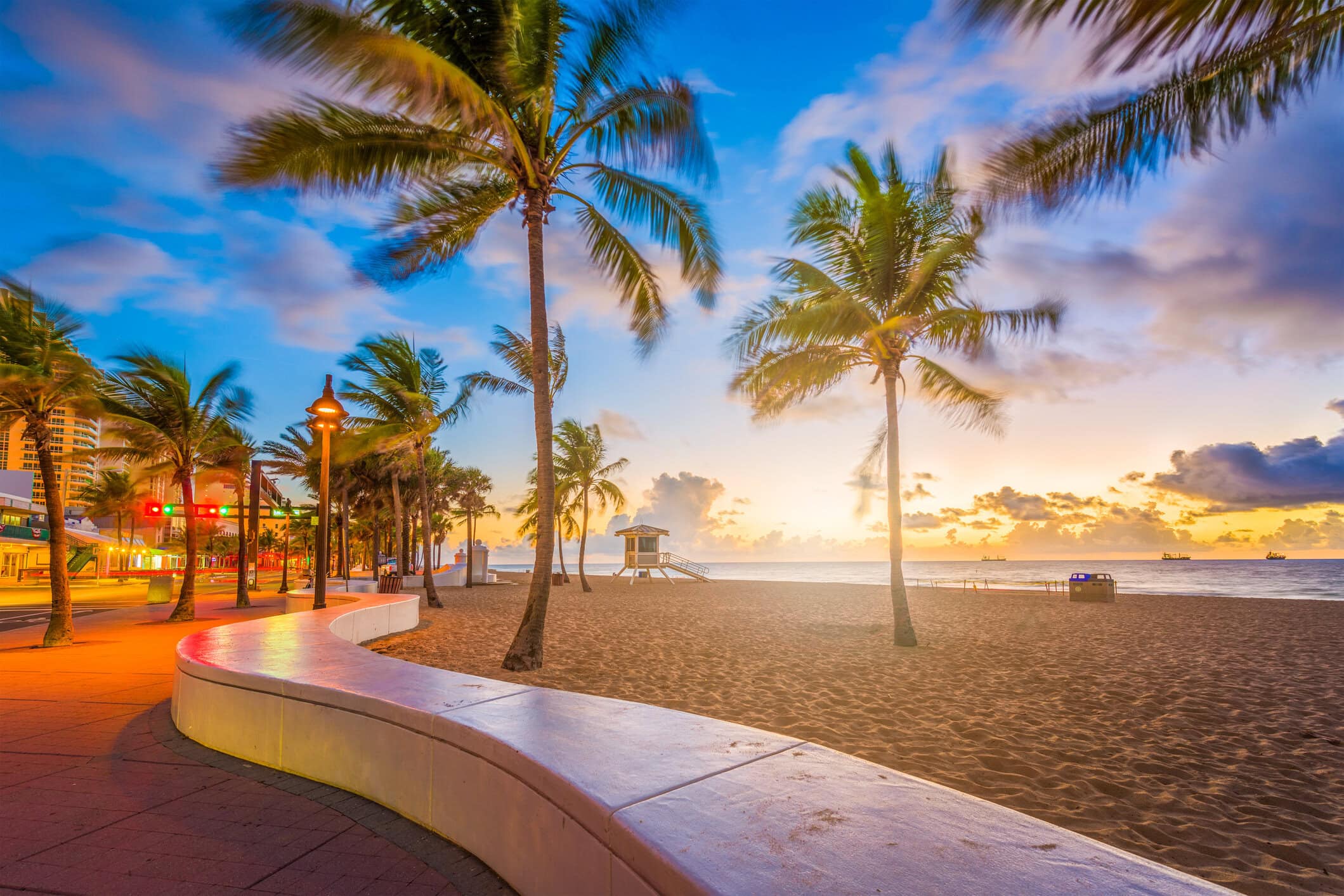 Sunrise over Fort Lauderdale Beach with palm trees, oceanfront promenade, sandy shoreline, and colorful morning sky.