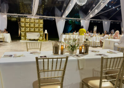 Glamorous 60th birthday dinner under clear tent with chandelier, gold chairs, and candlelit tables in Miami.