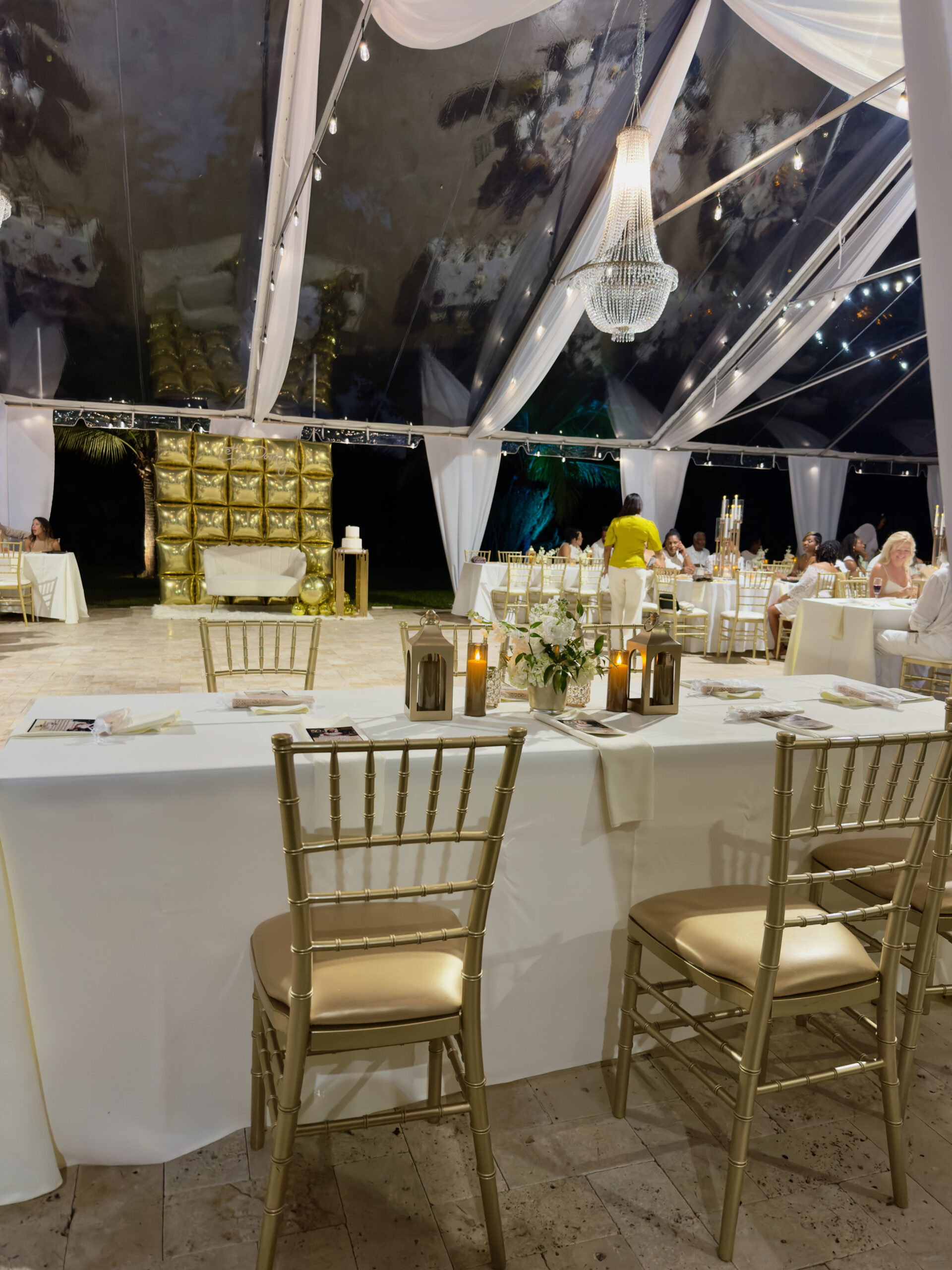 Glamorous 60th birthday dinner under clear tent with chandelier, gold chairs, and candlelit tables in Miami.