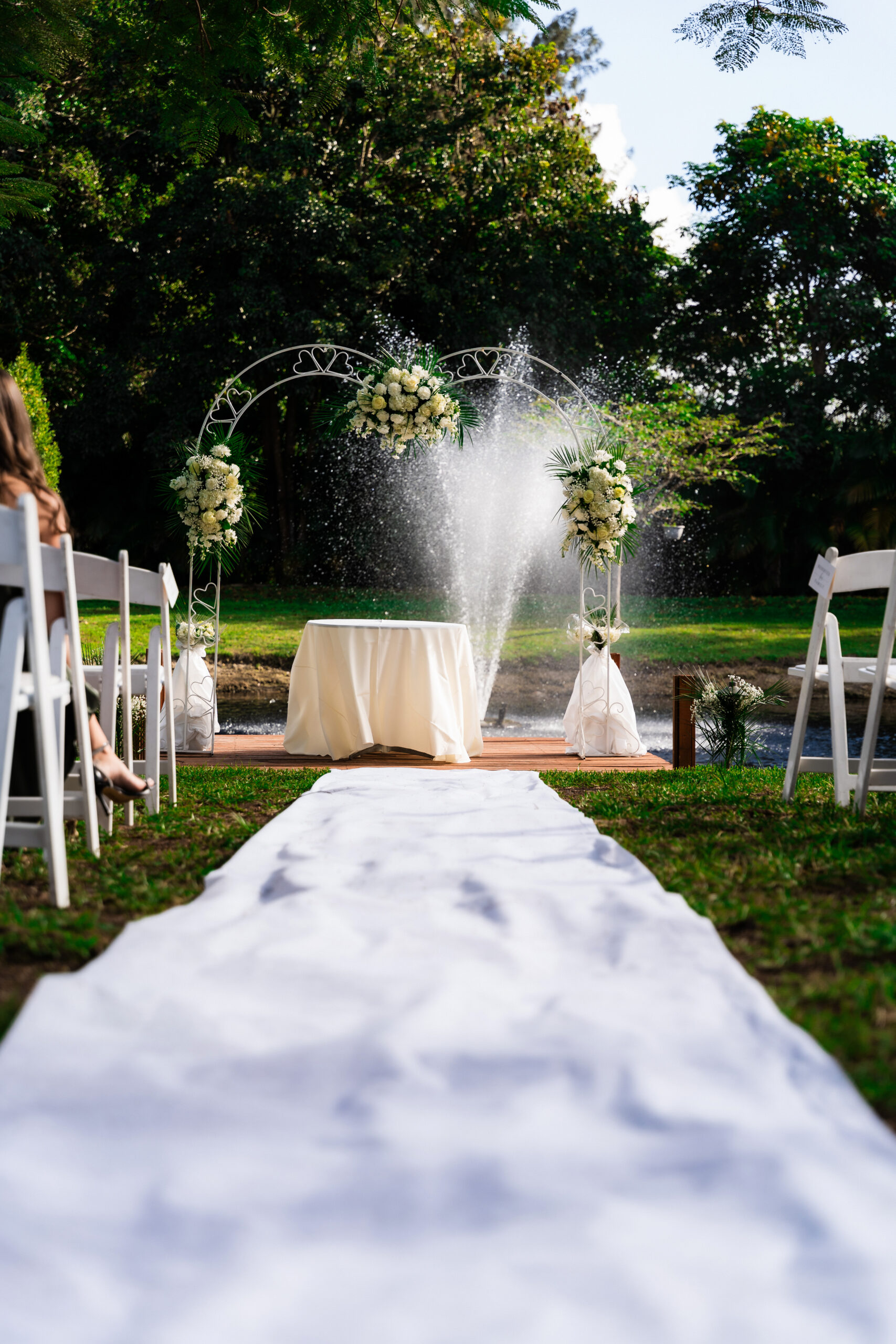 White aisle runner leading to floral arch and fountain backdrop at an outdoor wedding ceremony.
