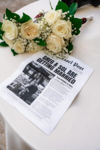 Newspaper-style wedding program with ivory rose bouquets on a table.