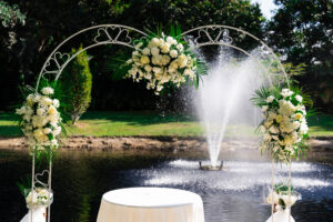 Elegant white floral ceremony arch with tropical greenery overlooking a pond and fountain at Saint Patrick Palace.