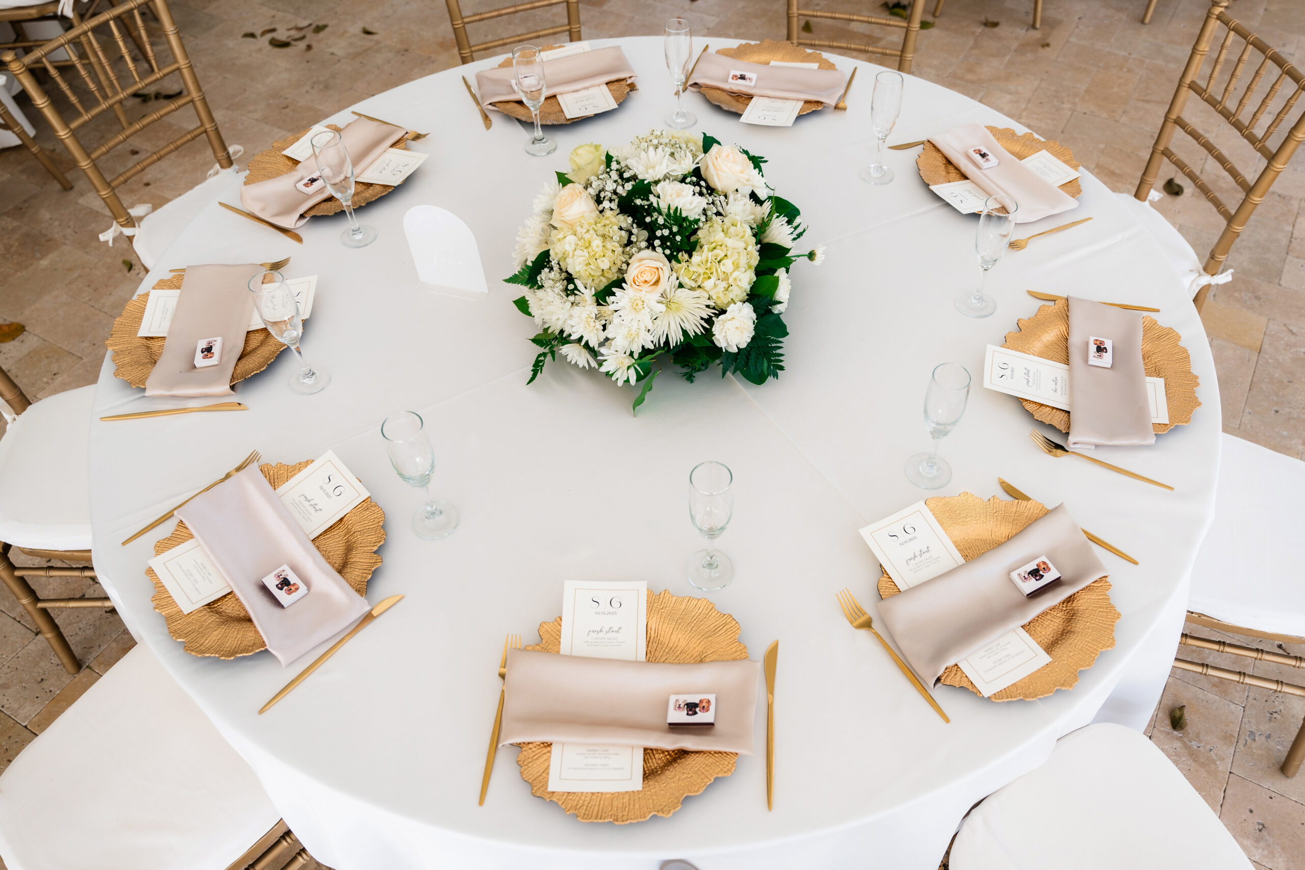 Round wedding table with gold chargers, champagne napkins, custom menus, and a white floral centerpiece.