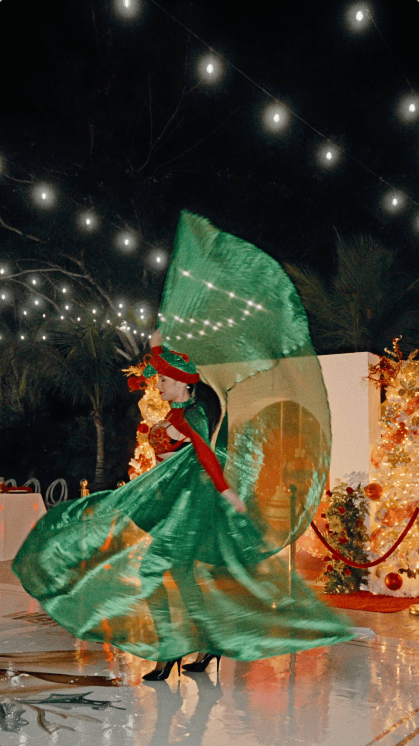 Holiday dancer twirling metallic green wings beside illuminated Christmas trees on an outdoor dance floor.