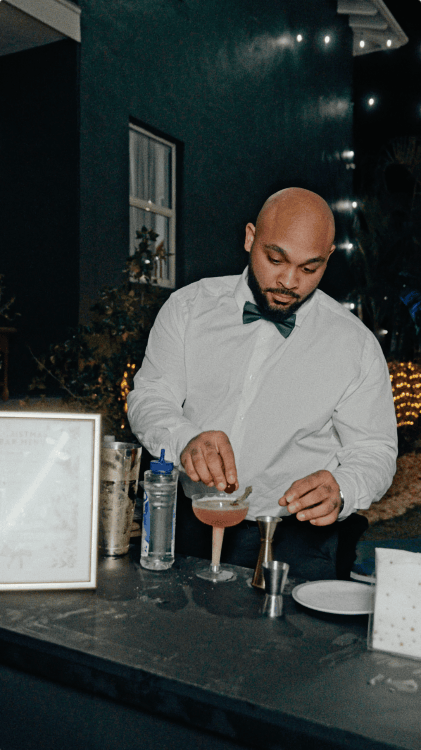 Bartender wearing a green bow tie garnishing a pink cocktail at an outdoor holiday party.