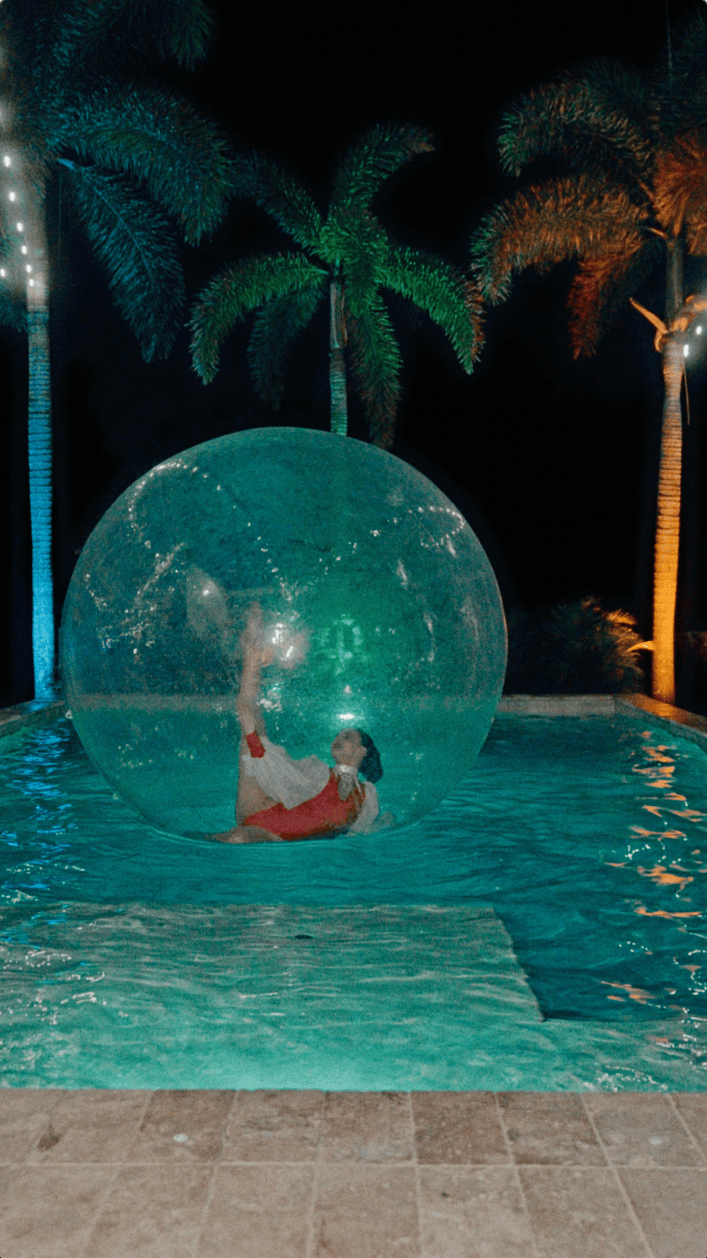 Holiday entertainer inside a transparent inflatable water ball floating in an illuminated outdoor swimming pool.