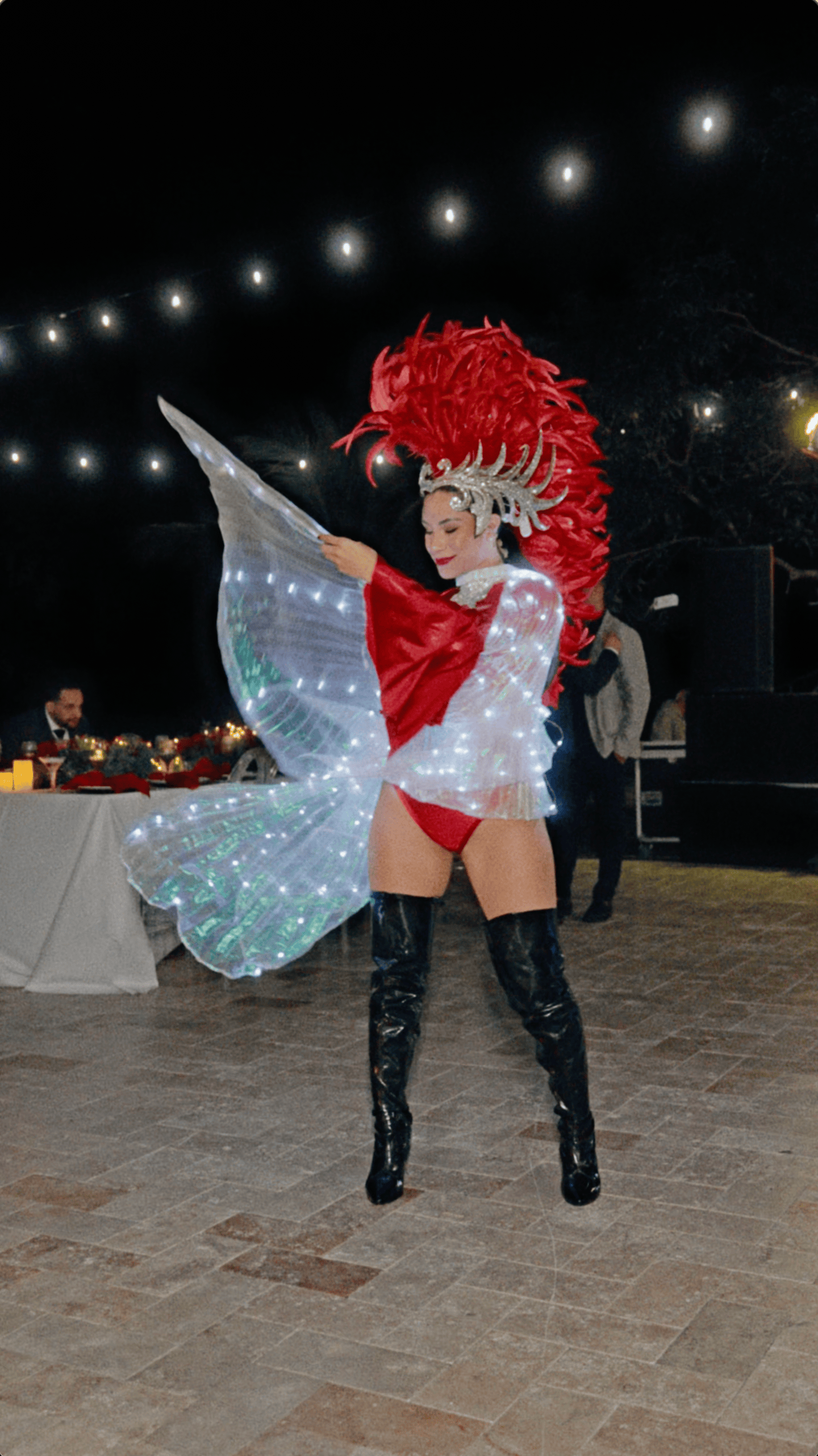 Christmas party performer in a red feathered headdress, black boots, and glowing wings beneath string lights.