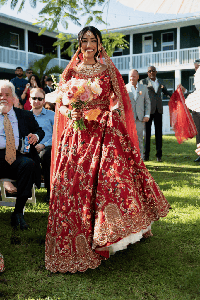 Indian bride in a red lehenga walking down the aisle during an outdoor wedding ceremony at Saint Patrick Palace in South Florida.