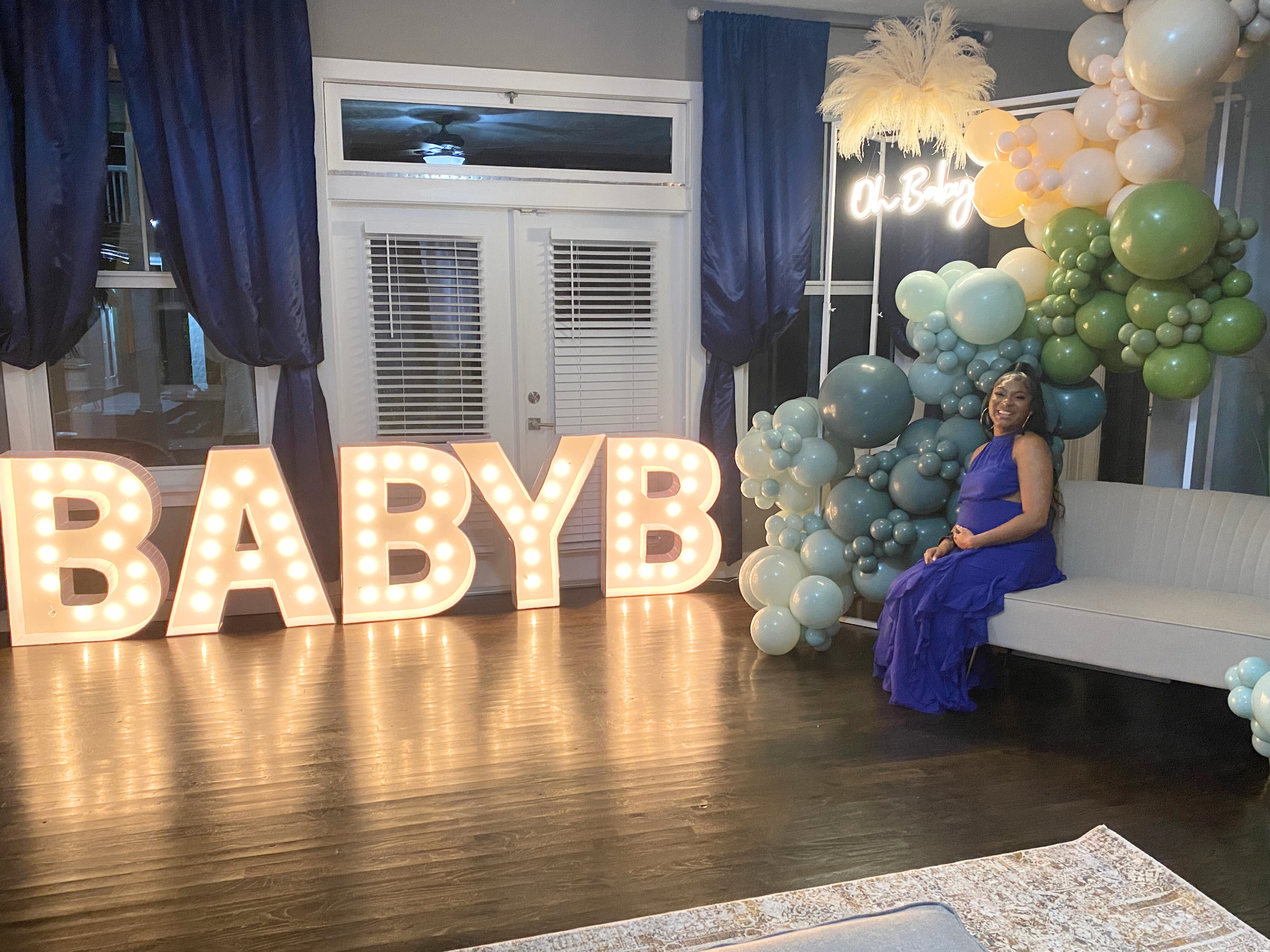 ndoor baby shower with marquee “BABY” lights, pastel balloon garland, and seated mom-to-be in blue dress
