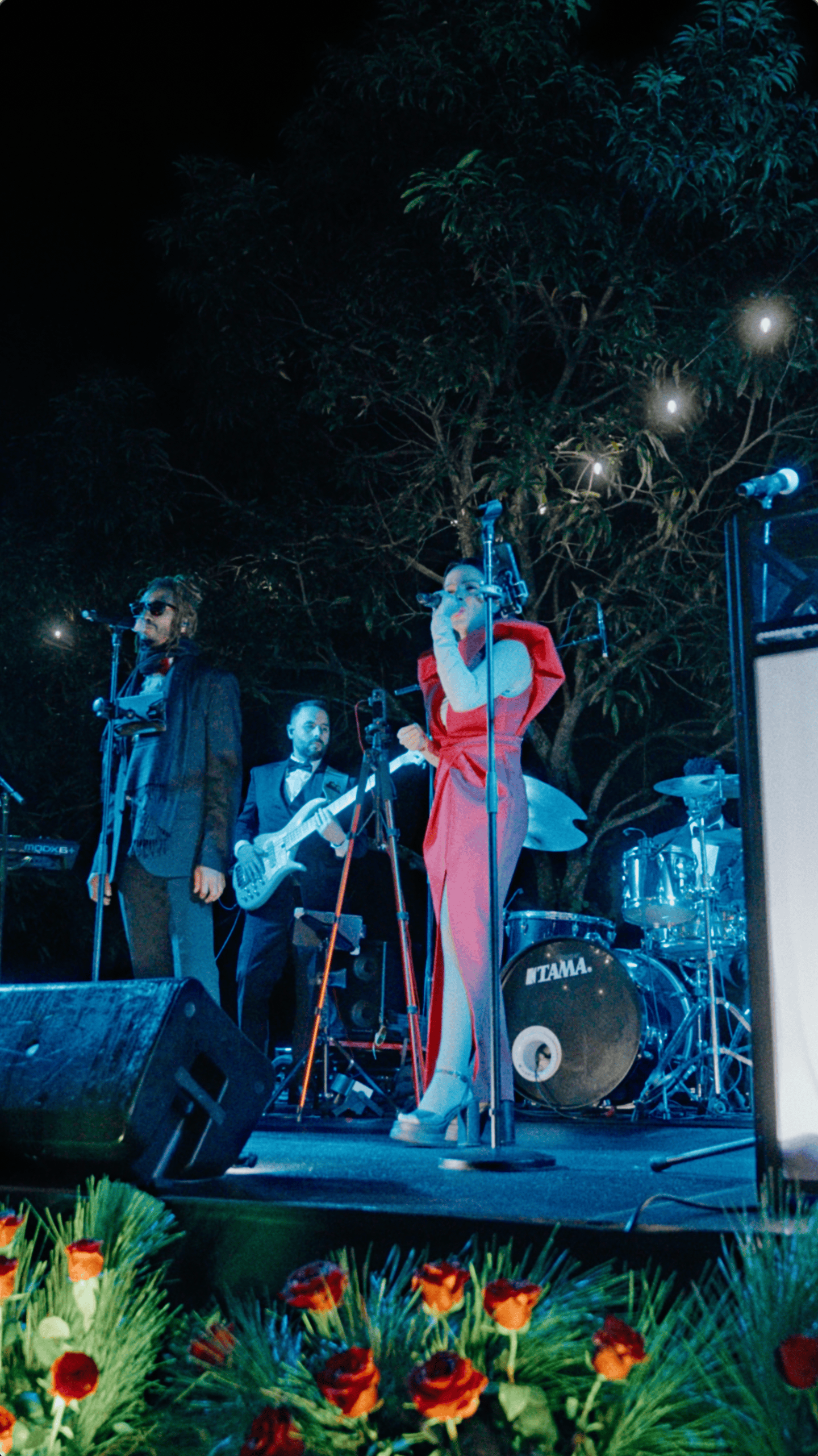 Female vocalist in a red gown performs with a live band on an outdoor stage beneath string lights.