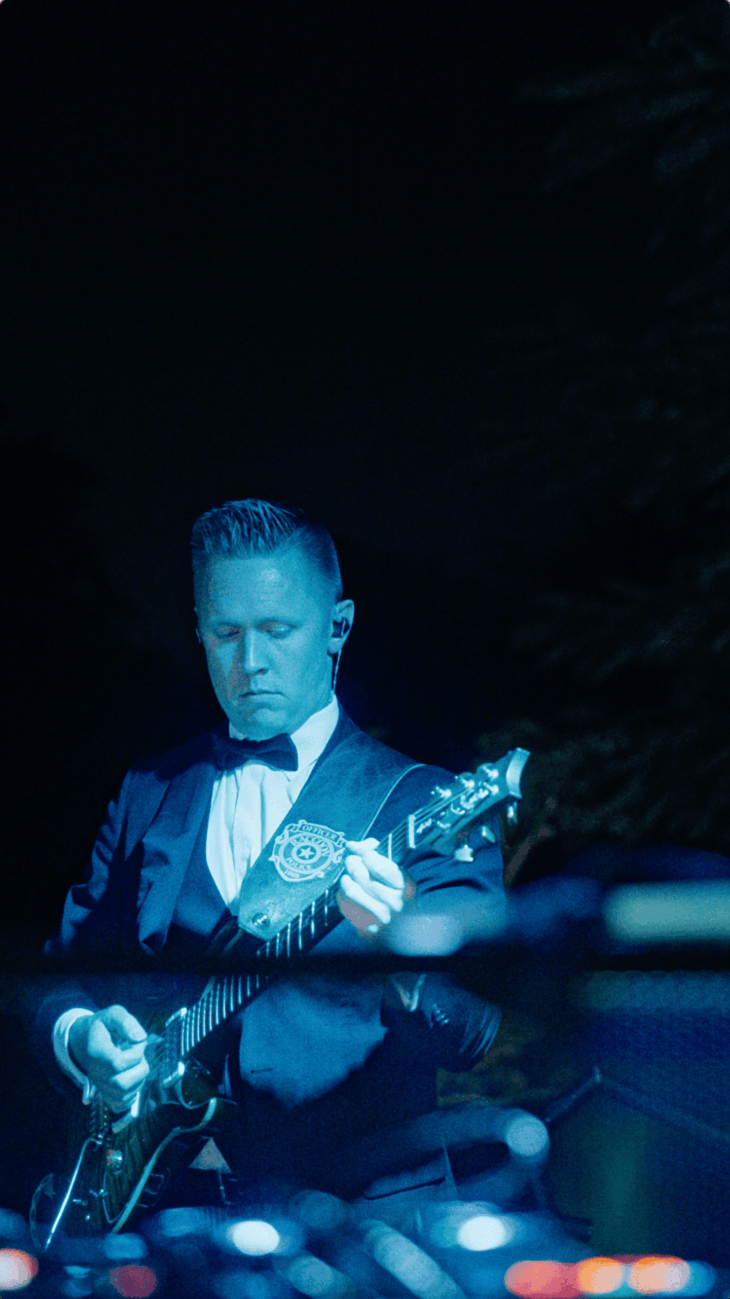 Guitarist in a dark suit and bow tie performs under blue stage lighting at an outdoor holiday event.