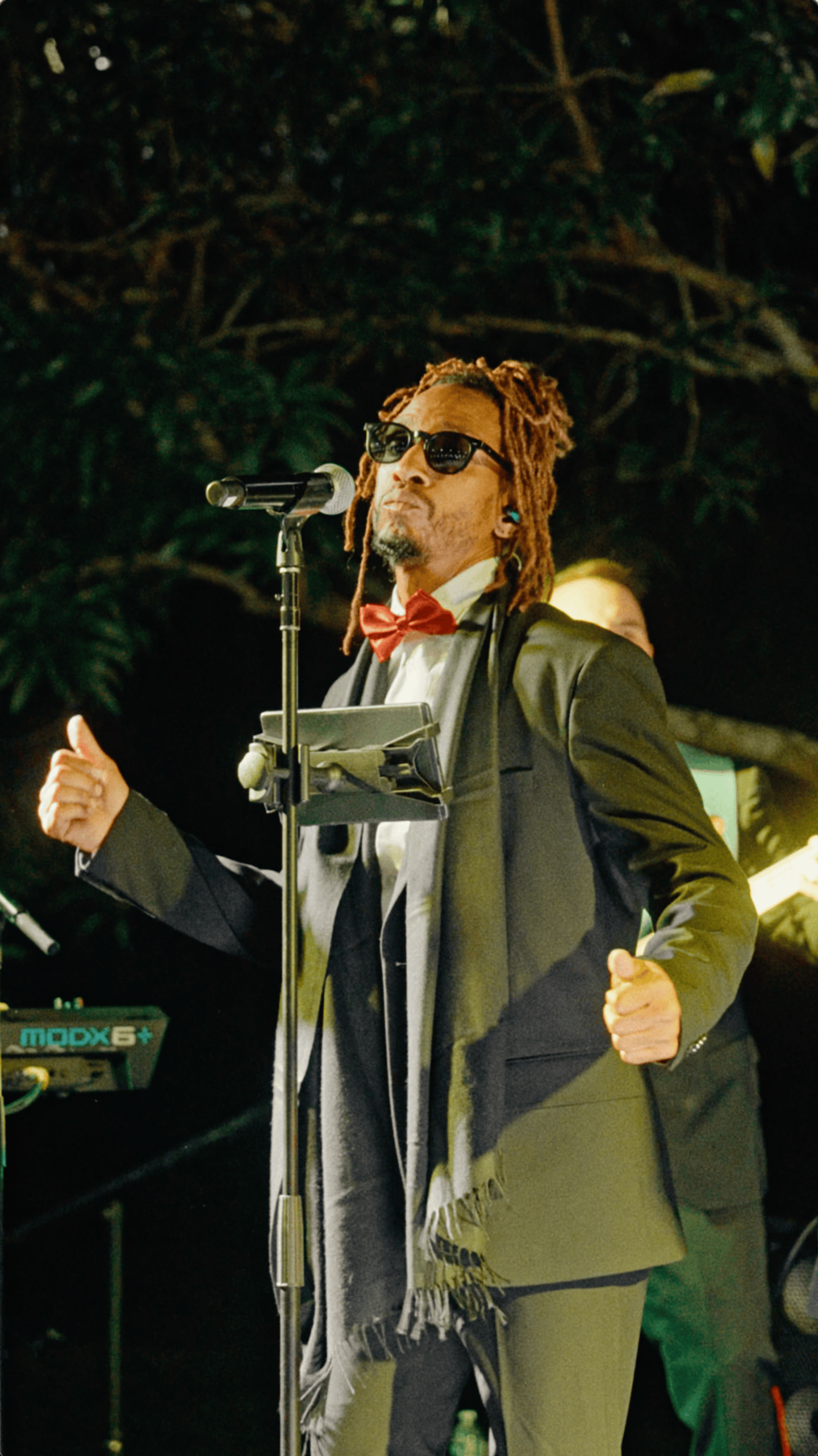 Male vocalist wearing a red bow tie and sunglasses performing at an outdoor holiday celebration.