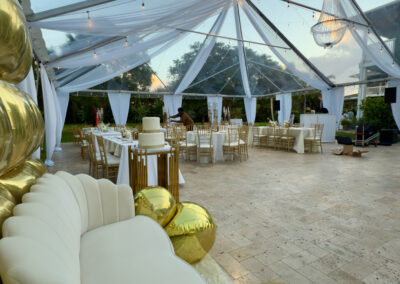 White lounge sofa and gold balloon décor under clear tent for elegant 60th birthday in Miami.