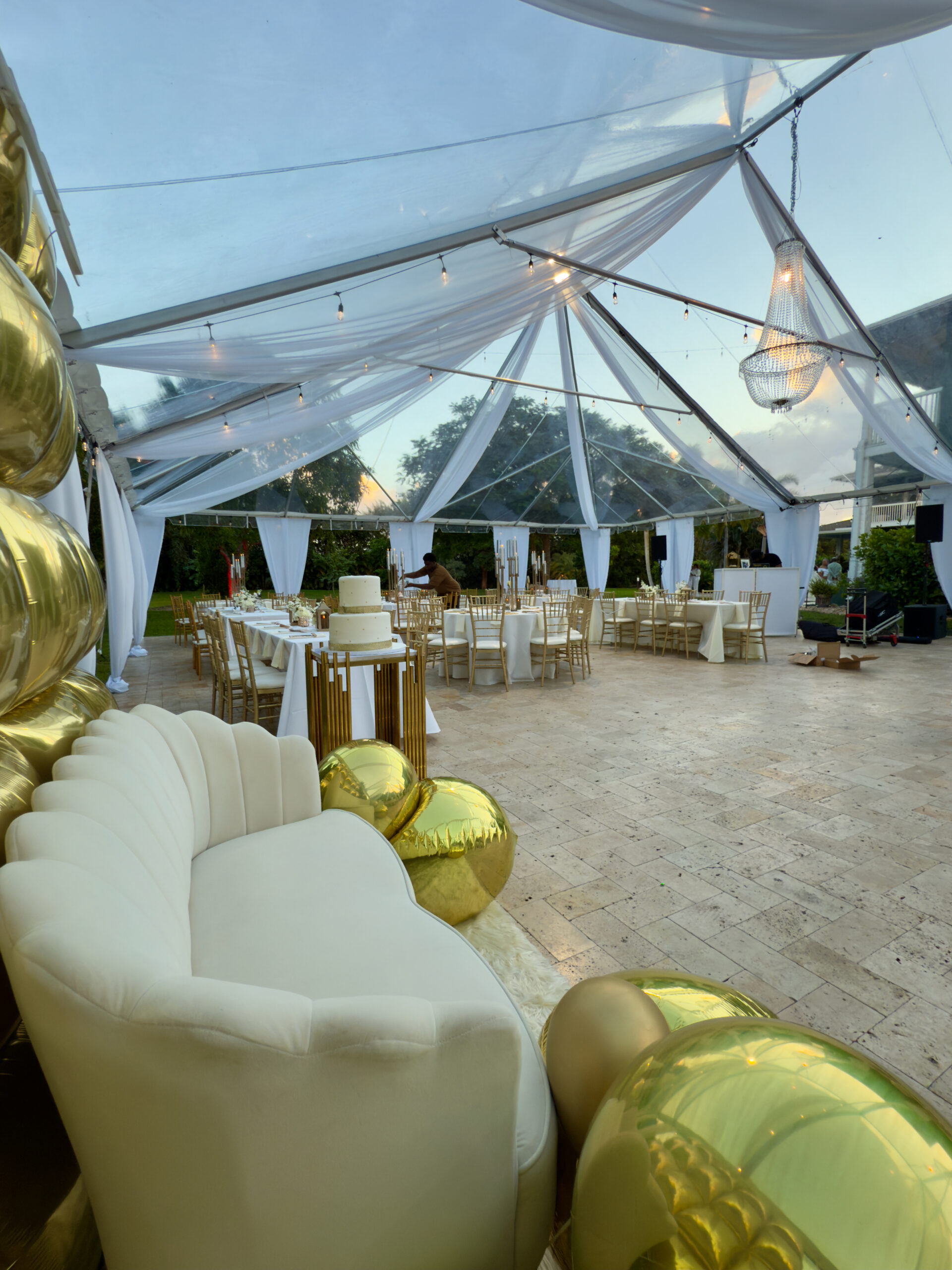 White lounge sofa and gold balloon décor under clear tent for elegant 60th birthday in Miami.
