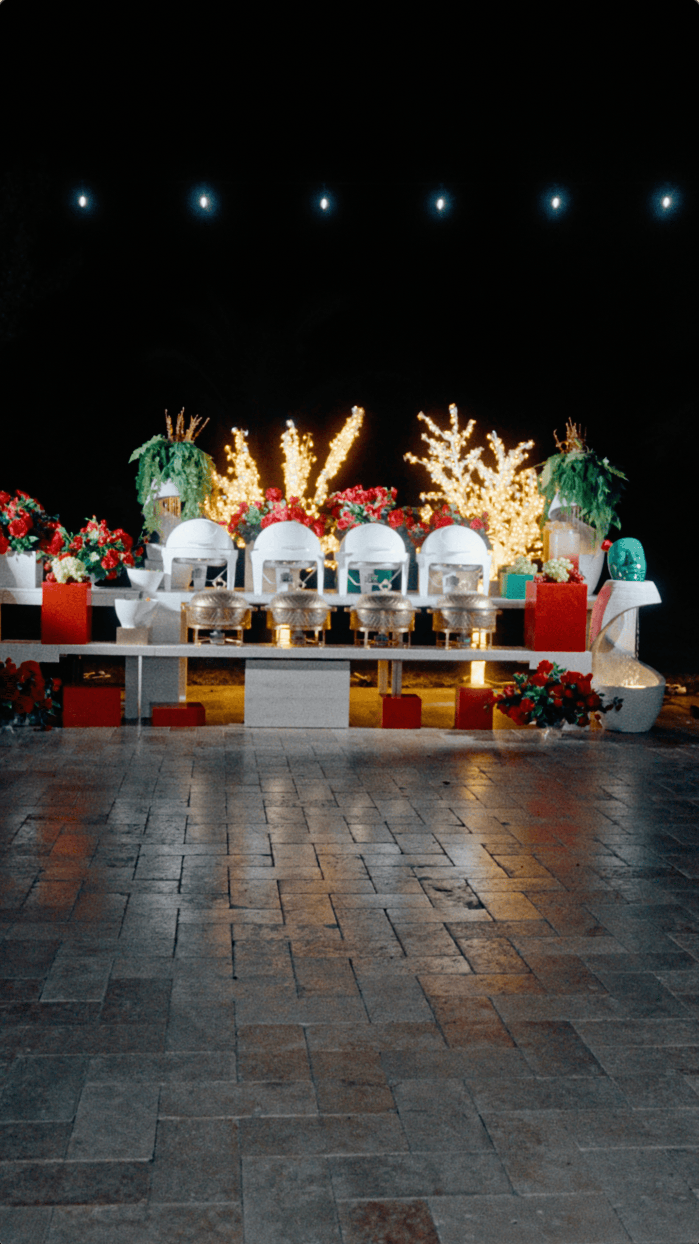 Luxury outdoor Christmas buffet with white serving stations, gold chafing dishes, red flowers, and illuminated trees.