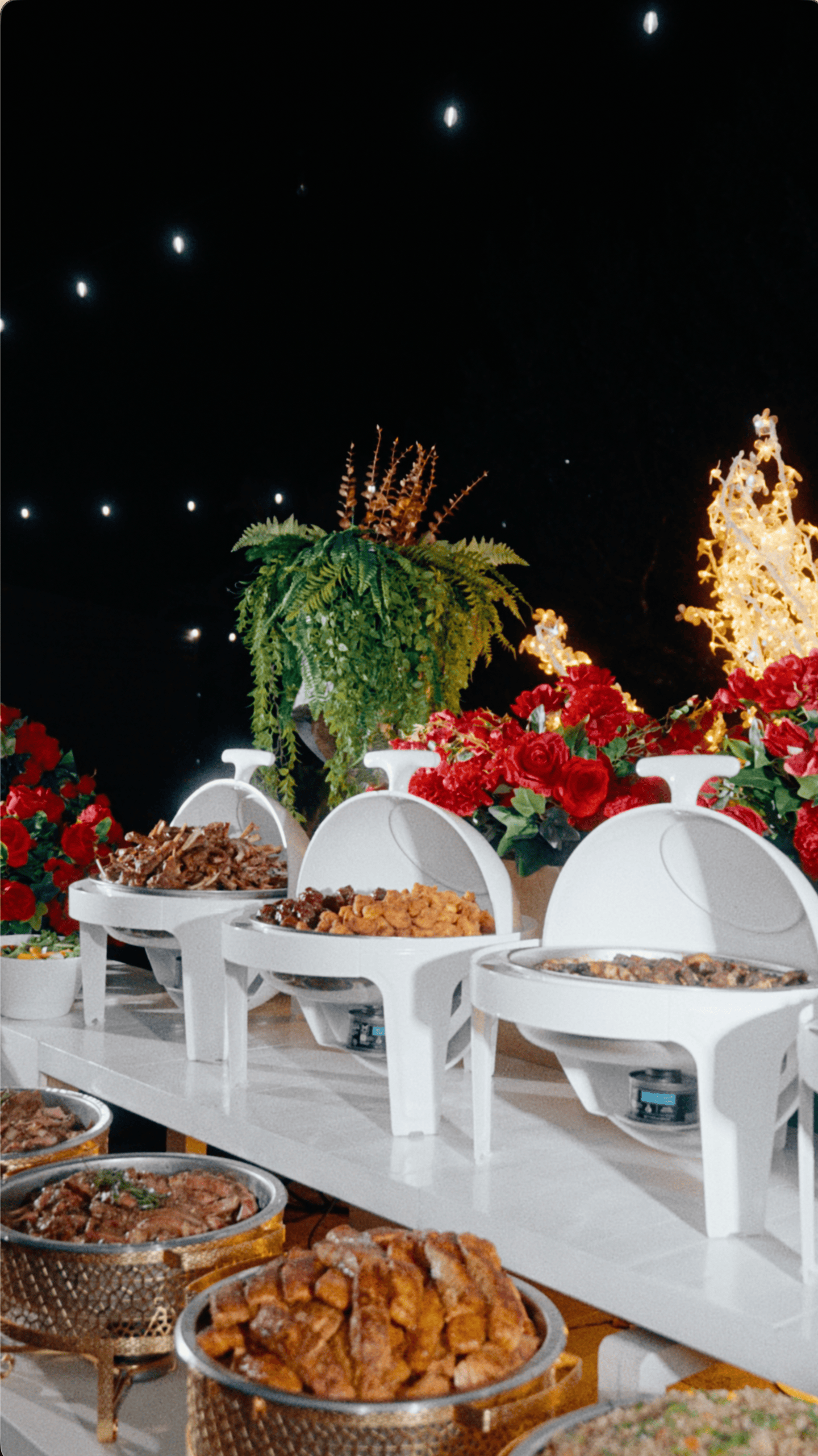 Elegant holiday buffet with white chafing dishes, gold serving stands, red flowers, and illuminated decorations.