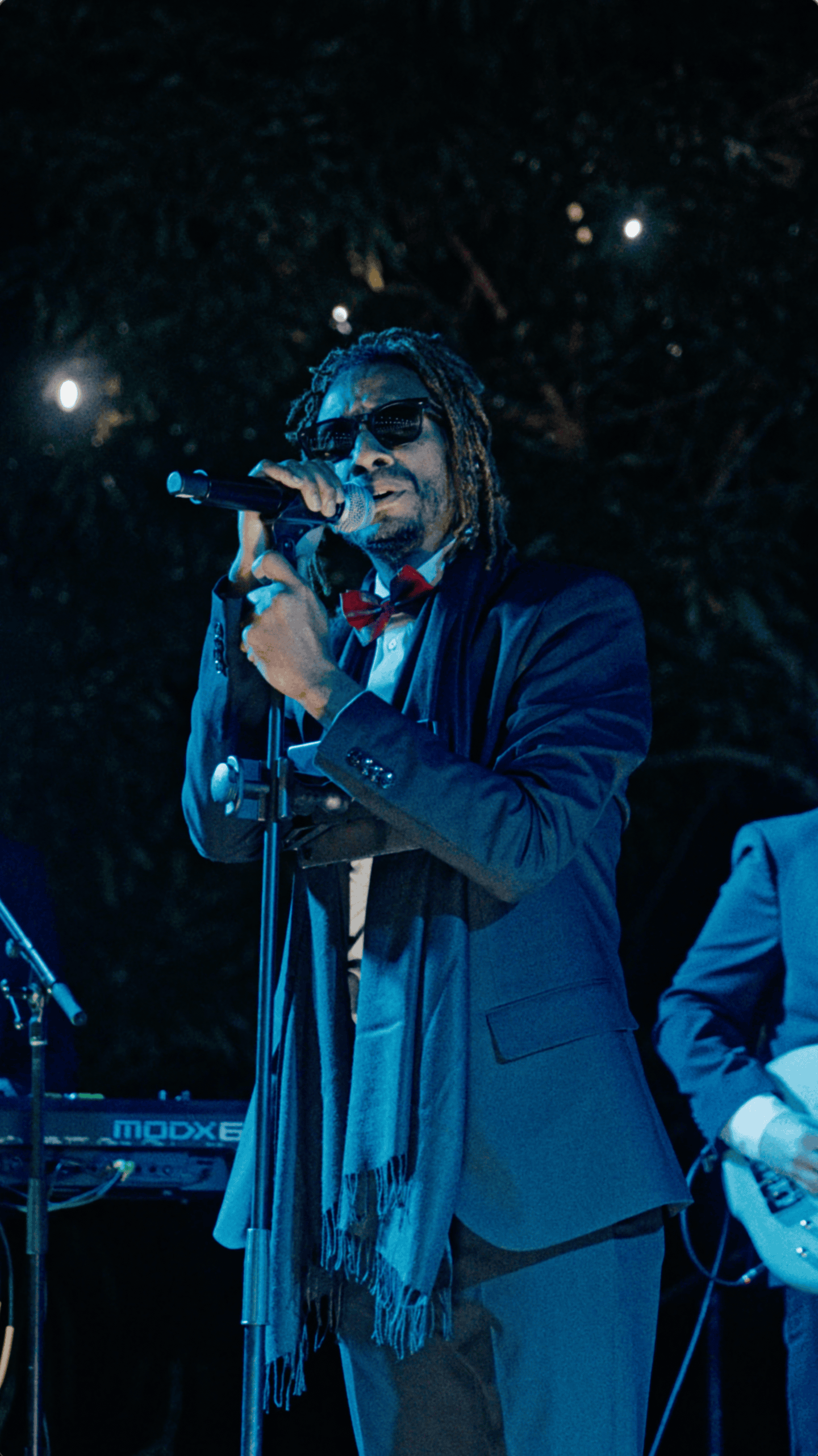 Male vocalist wearing sunglasses and a dark suit sings into a microphone during an outdoor holiday celebration.