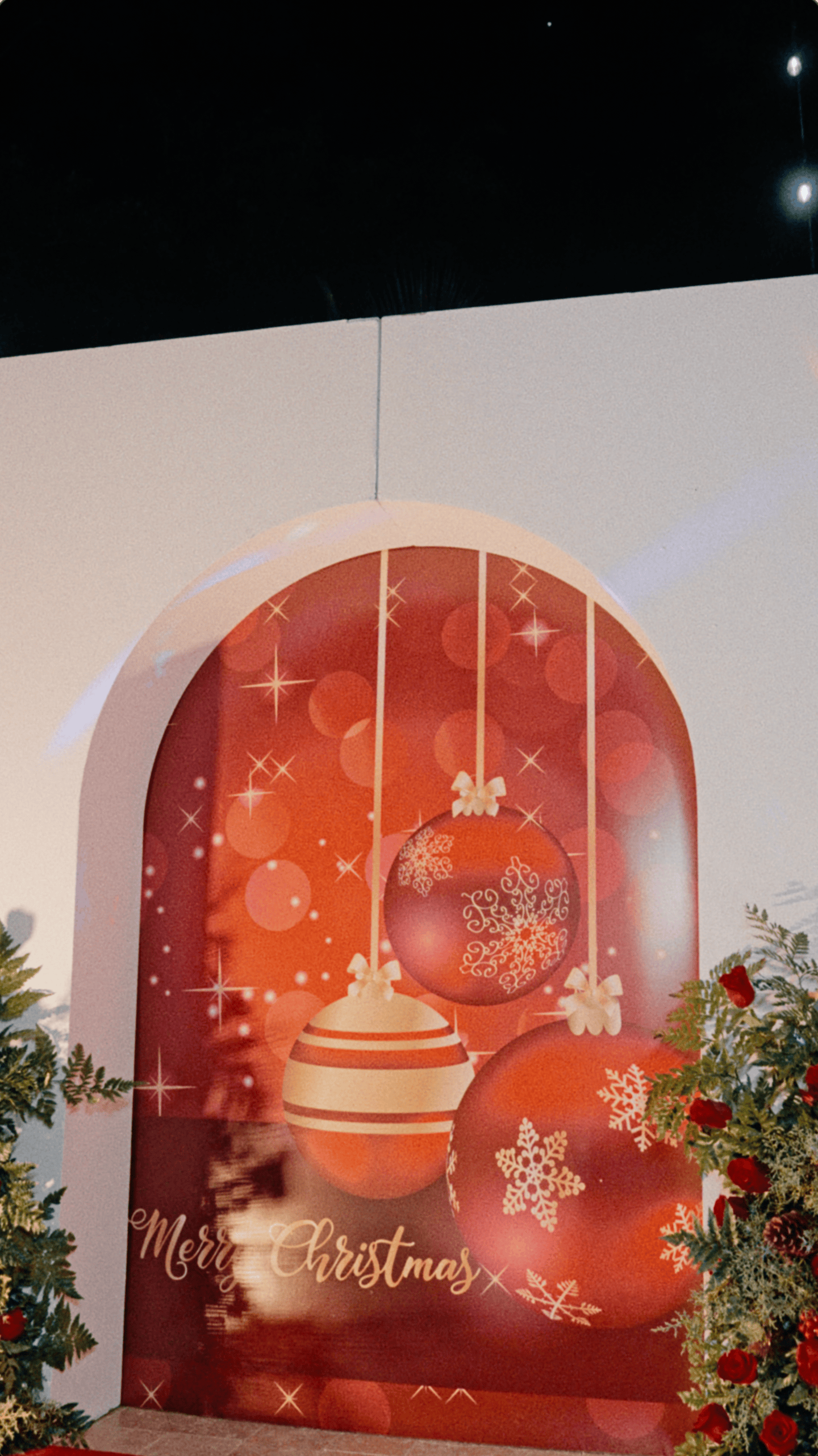 Merry Christmas photo wall with oversized red ornaments, white arch, red roses, and holiday greenery.