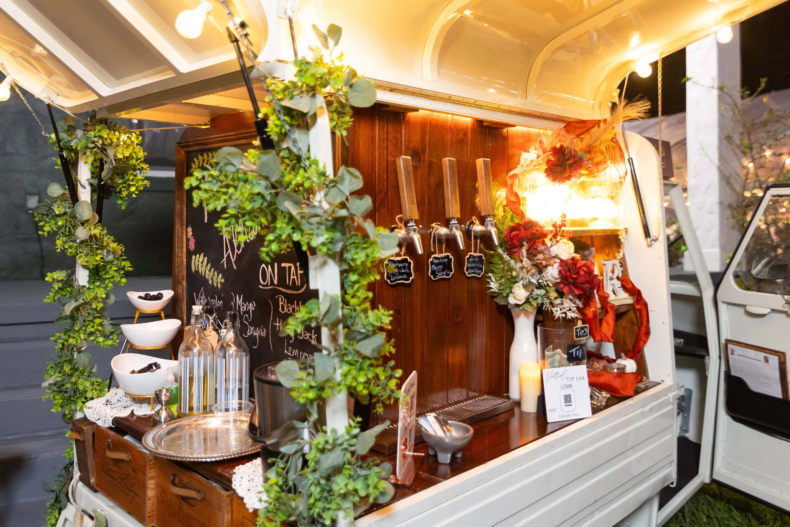 Vintage mobile wedding bar with custom cocktails, greenery decor, and chalkboard menu at outdoor reception.