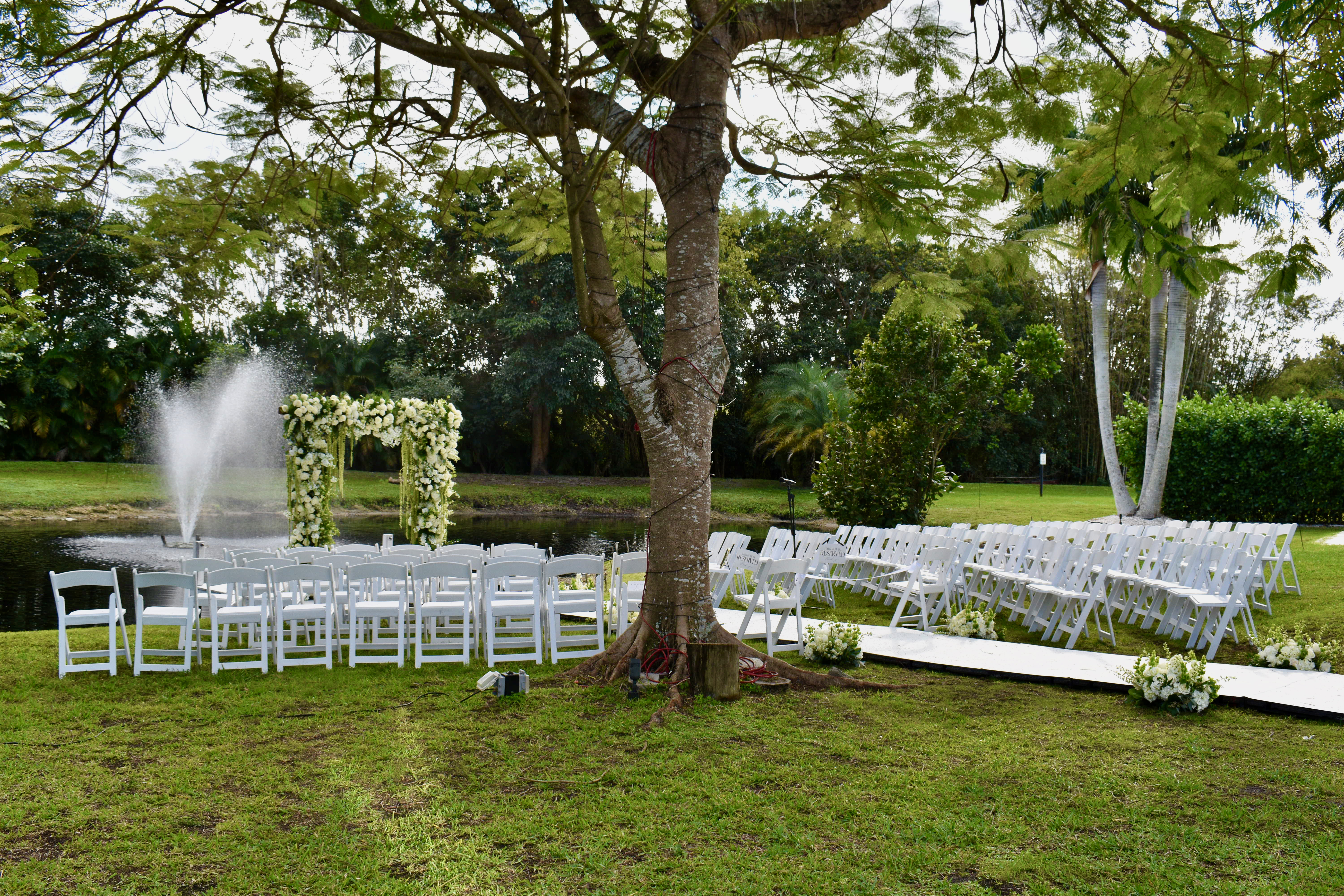 Outdoor estate wedding ceremony seating with floral arch beside pond in garden setting