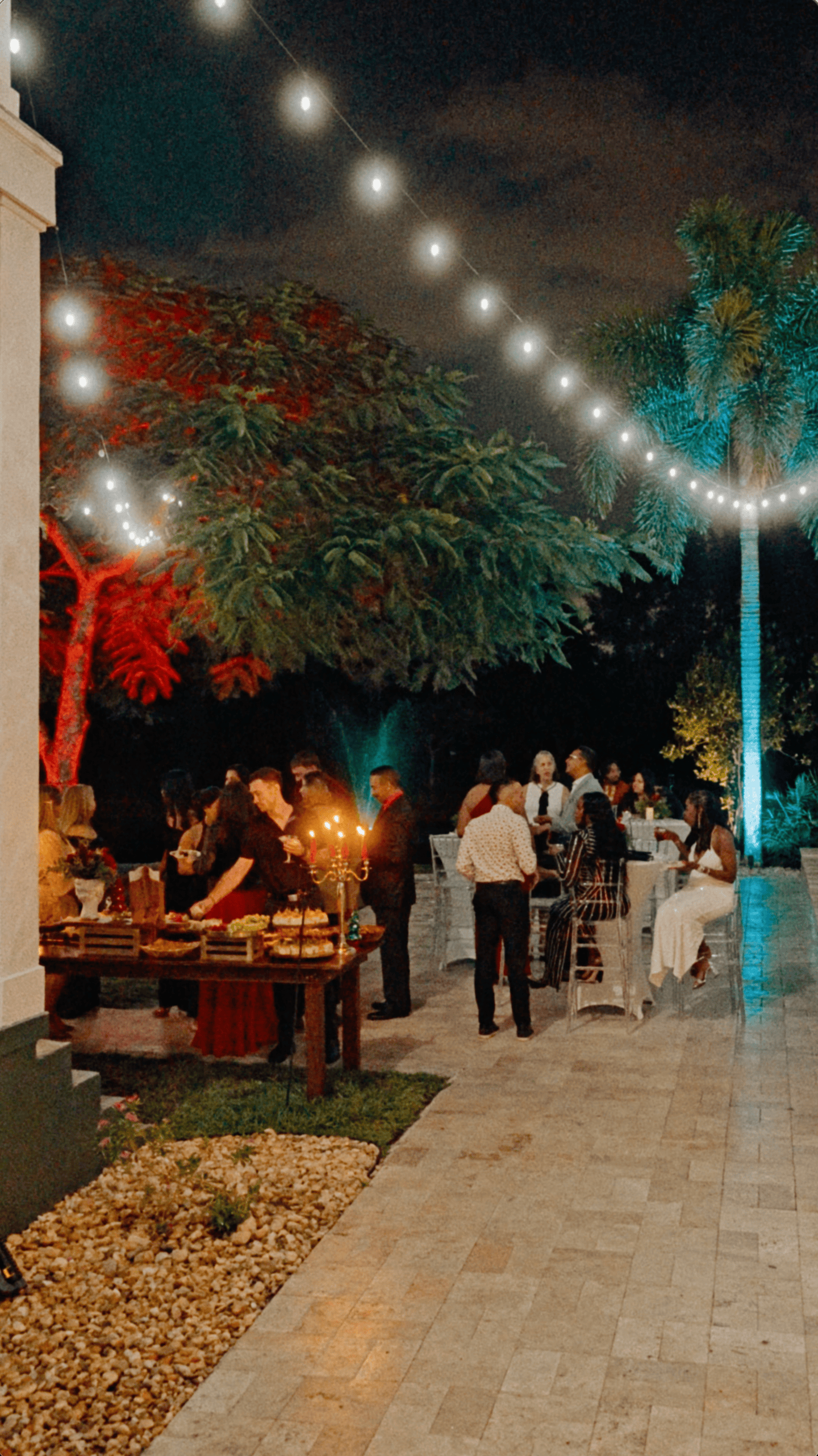 Outdoor holiday reception with guests, catering tables, bistro lights, and colorful landscape lighting.