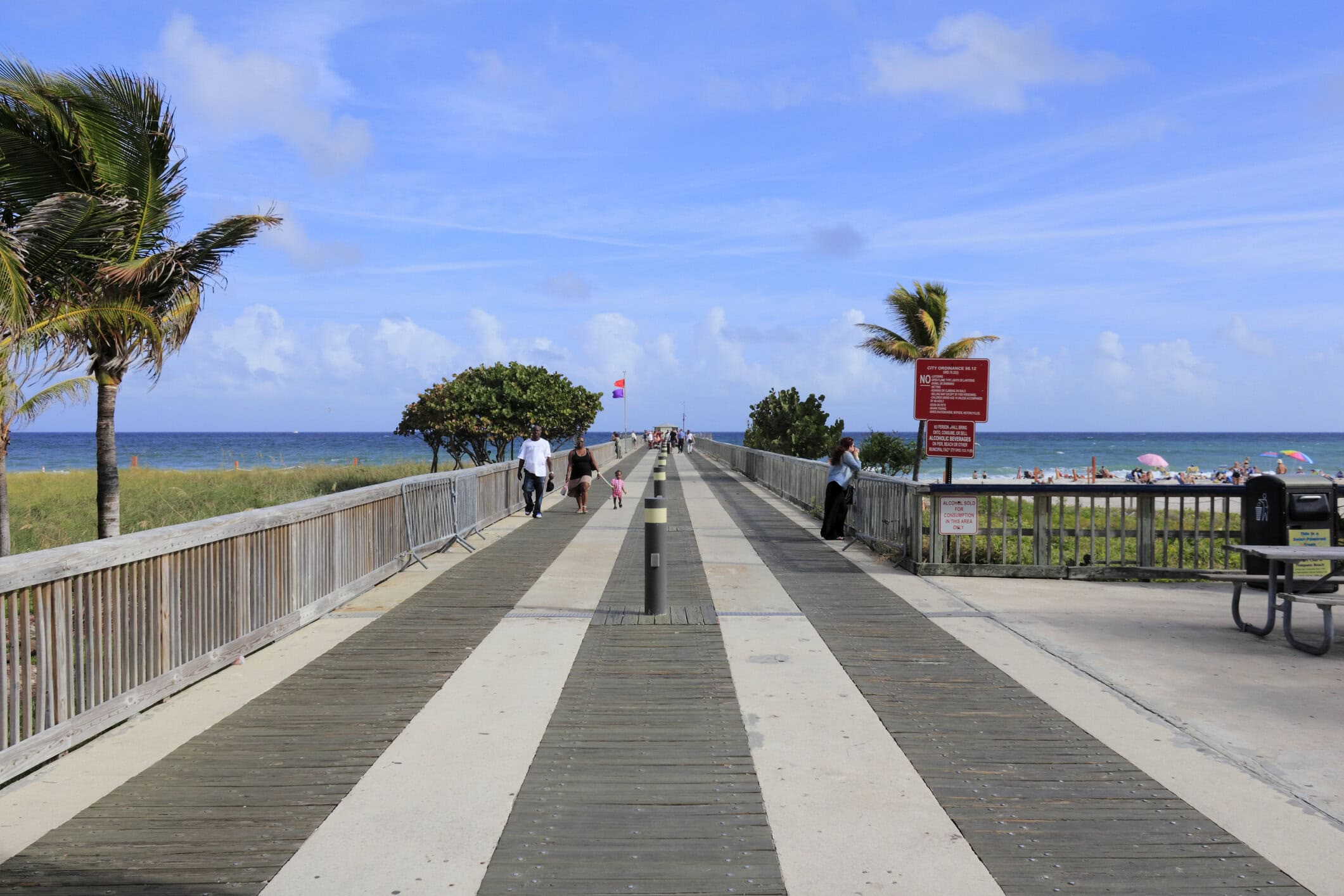 Historic Pompano Beach pier walkway with visitors, palm trees, sandy shoreline, and Atlantic Ocean views.