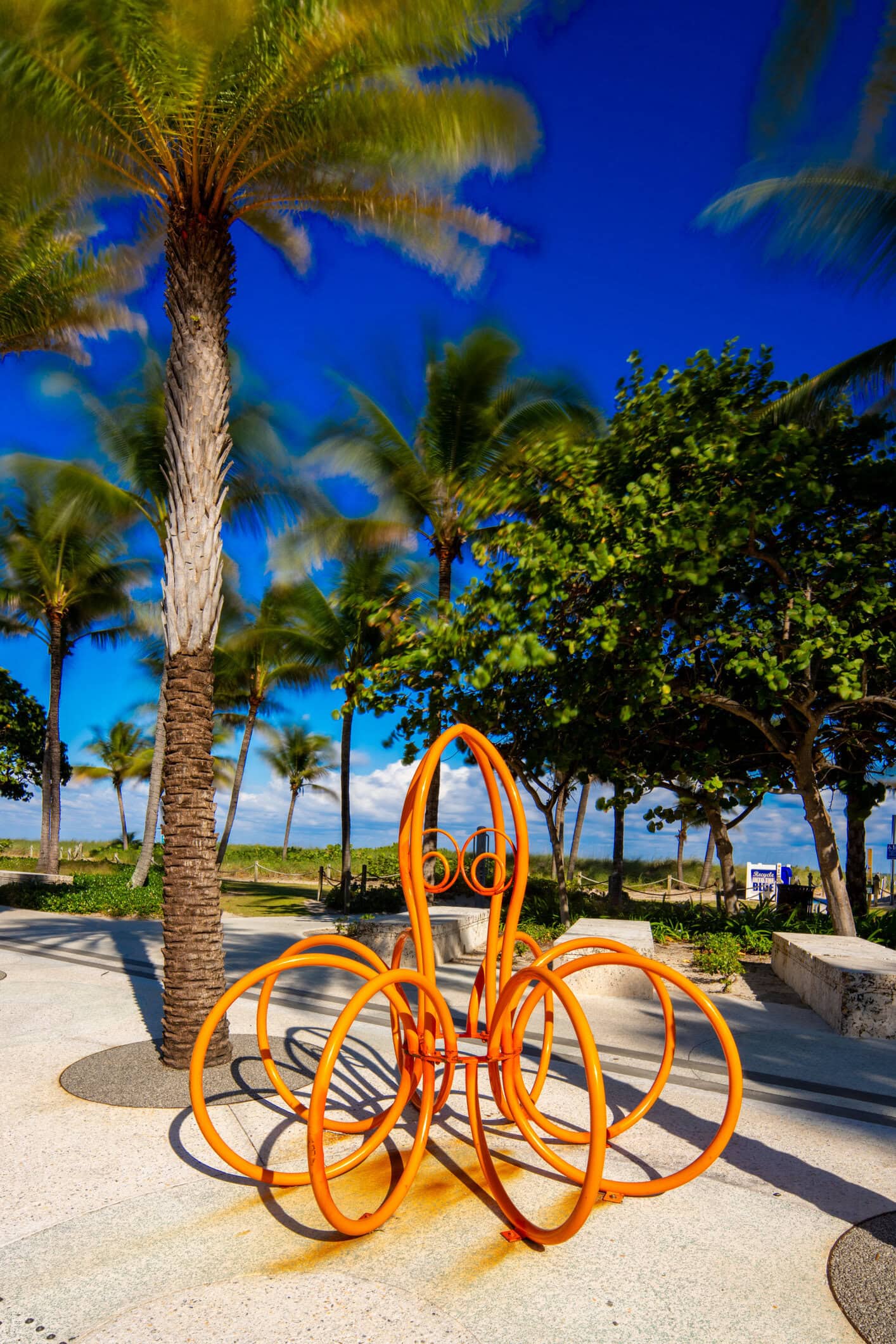 Bright orange bicycle-inspired sculpture surrounded by palm trees and tropical landscaping near Pompano Beach.