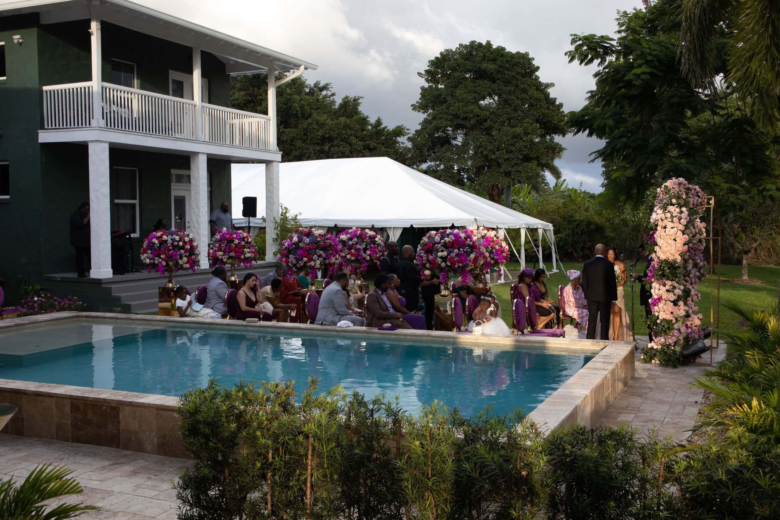 Poolside outdoor wedding ceremony with colorful floral arrangements, guest seating, and a nearby white reception tent in Davie.