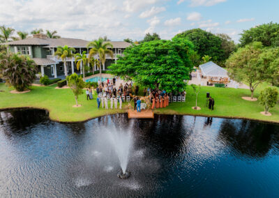 Aerial view of a lakeside wedding ceremony at Saint Patrick Palace in Davie, South Florida, with a fountain, pool, and luxury reception tent.