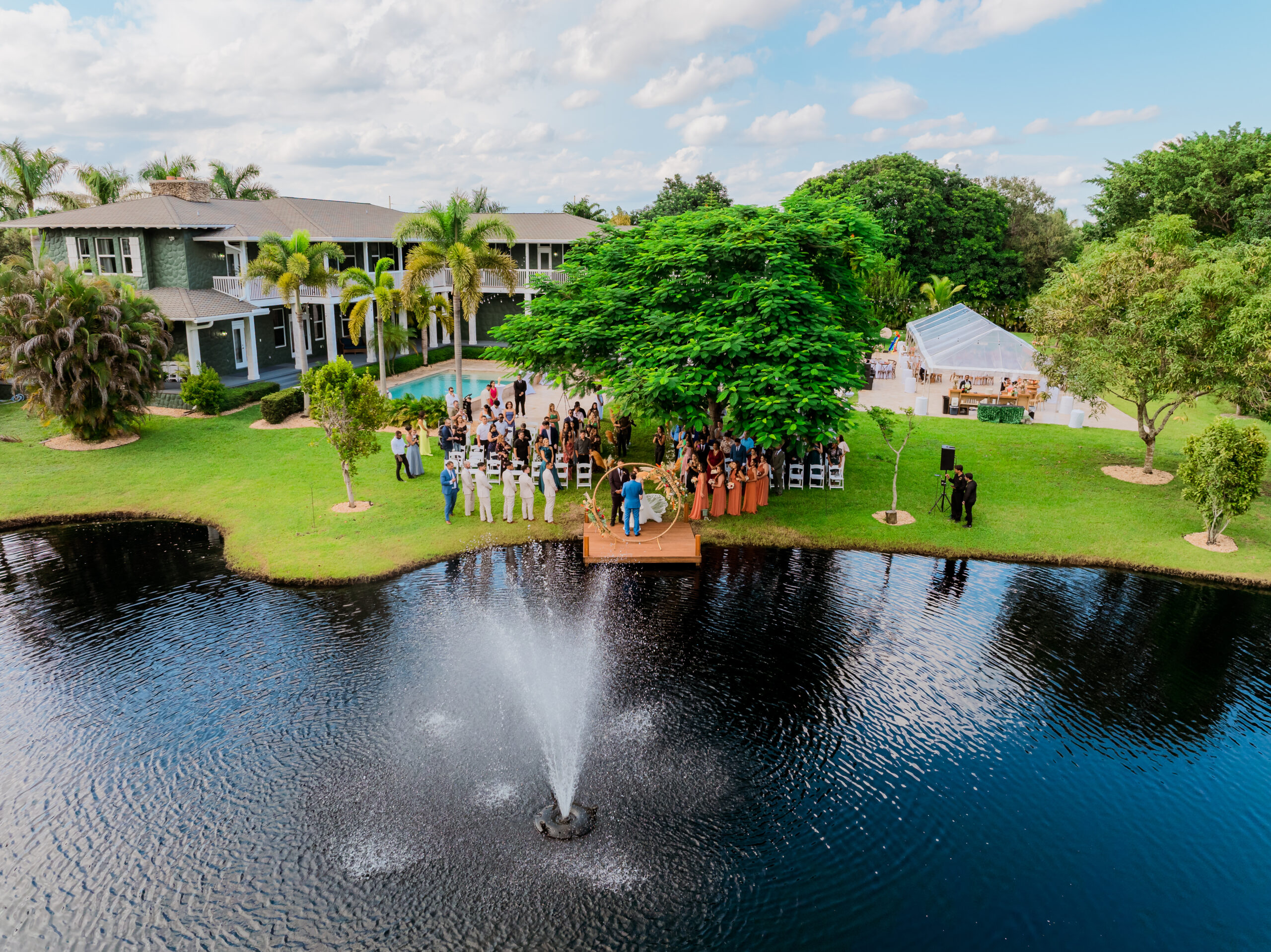 Aerial view of a lakeside wedding ceremony at Saint Patrick Palace in Davie, South Florida, with a fountain, pool, and luxury reception tent.