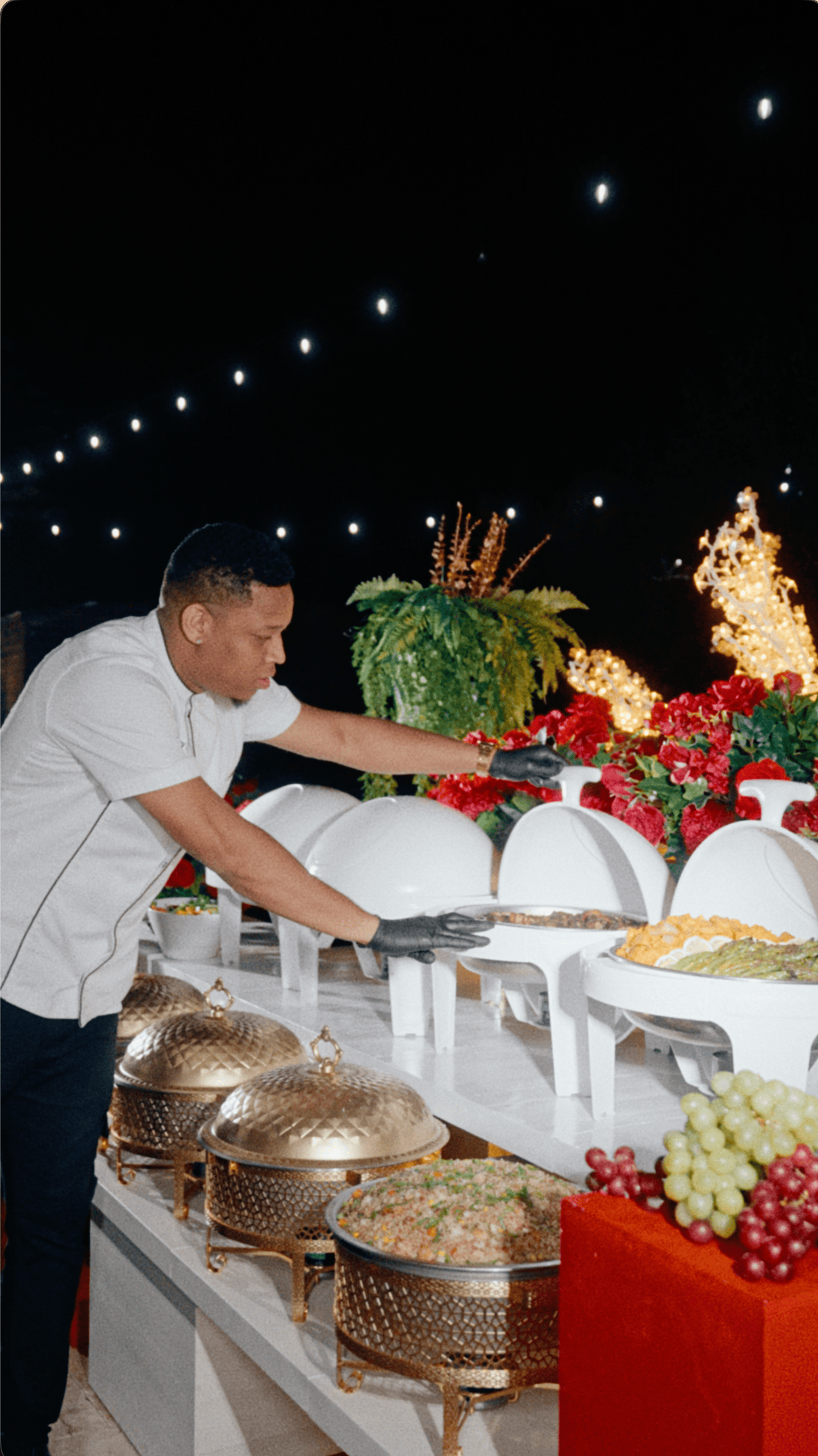 Catering staff member arranging an outdoor holiday buffet with white chafing dishes and festive red flowers.