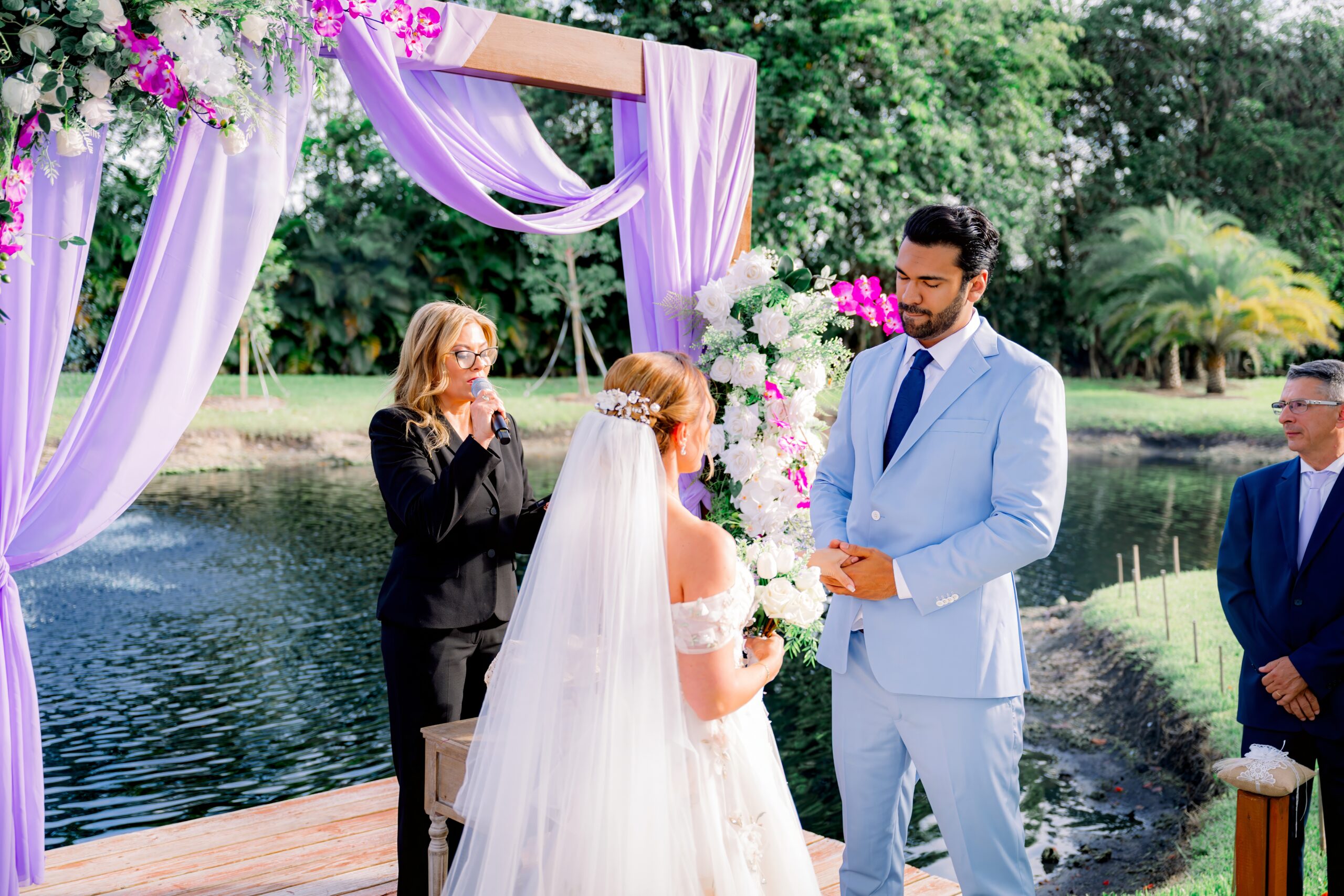 Couple exchanging vows by a lakeside dock under a draped purple floral arch at Saint Patrick Palace in Davie, Florida.
